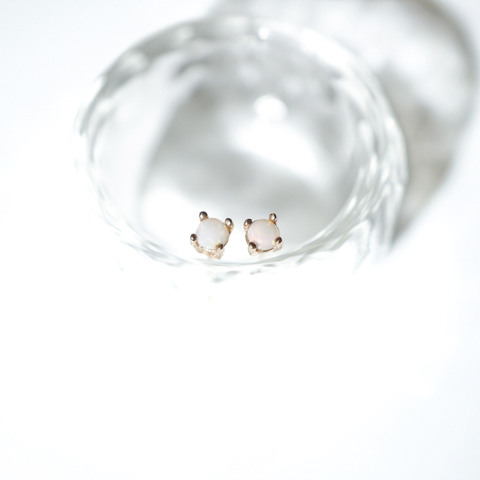 A pair of small gold stud earrings with oval pale pink stones, displayed inside a clear glass dish on a bright white background.