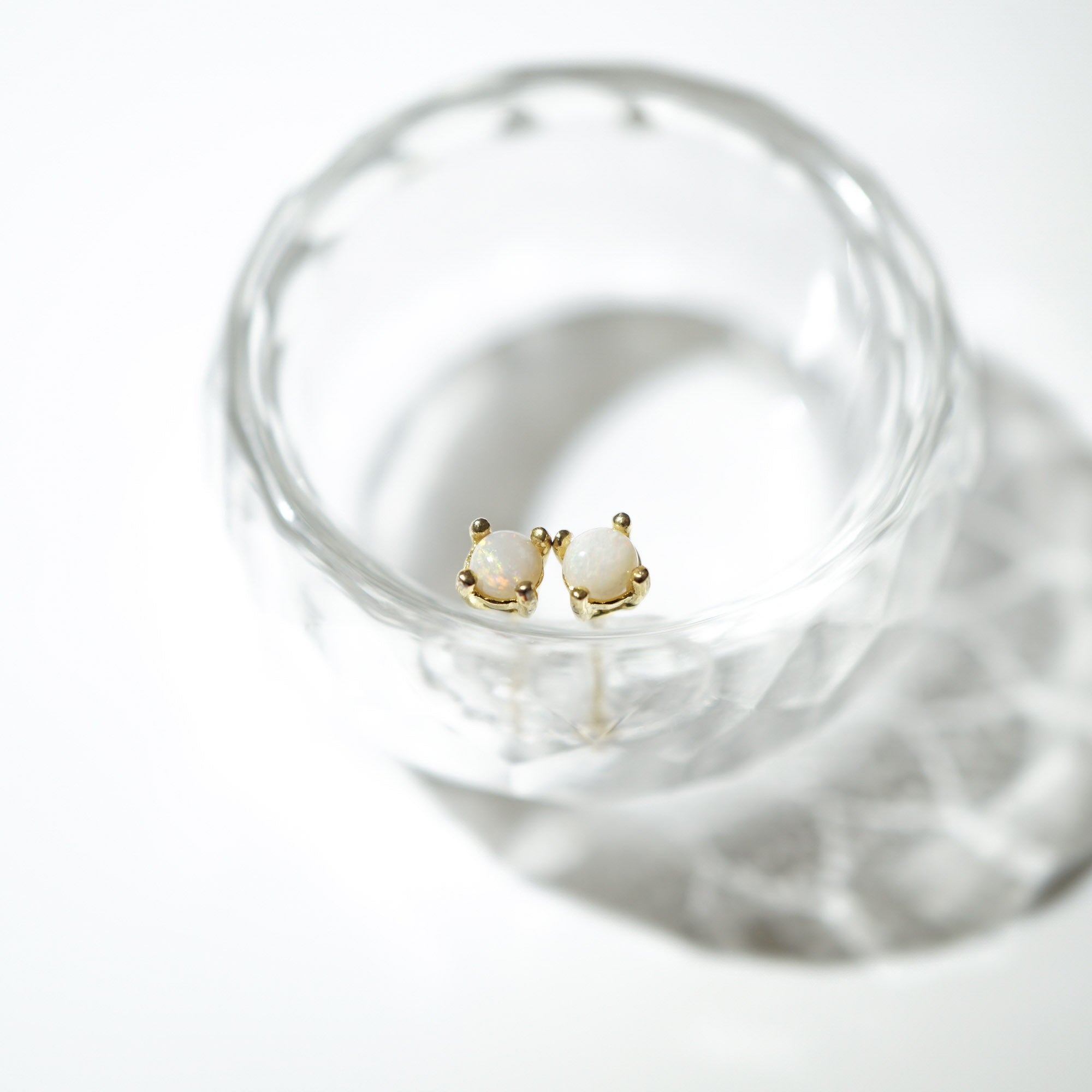 A pair of small gold stud earrings with oval white stones, displayed inside a clear glass bowl on a bright white background.