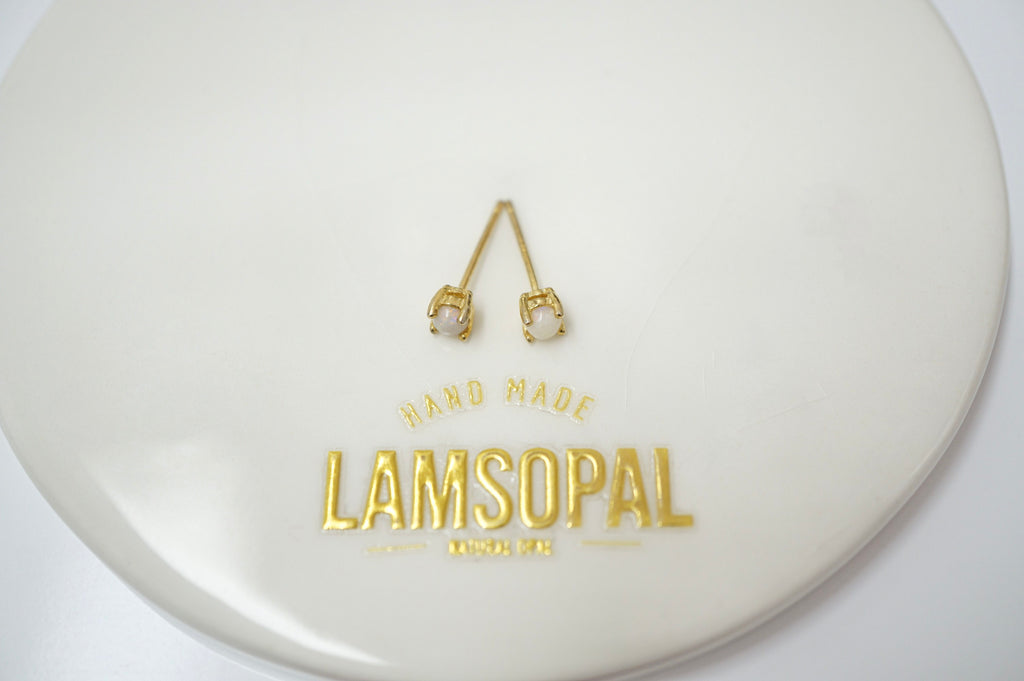 A pair of gold-toned stud earrings with clear rectangular gemstones, displayed on a glossy white circular surface with the brand name Lamsopal printed in gold below.