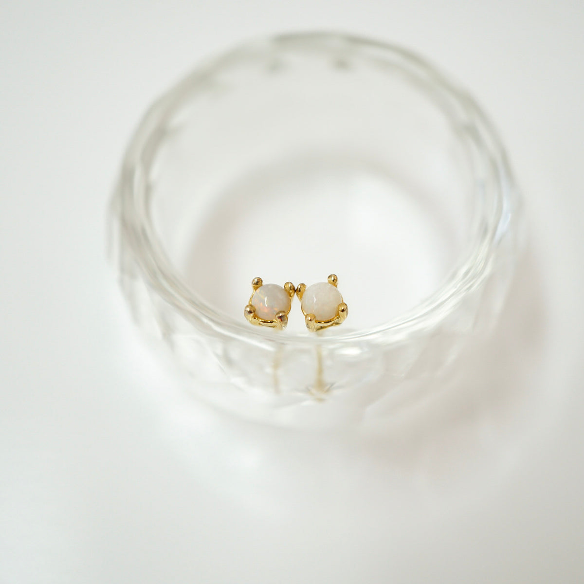 A pair of gold stud earrings with oval milky white stones, displayed inside a clear textured glass bowl on a white background.