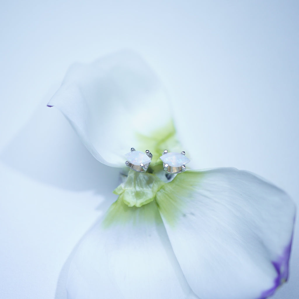 A pair of silver stud earrings with white cabochon stones, delicately resting on the center of a white flower with a soft green and purple center, set against a clean white background.