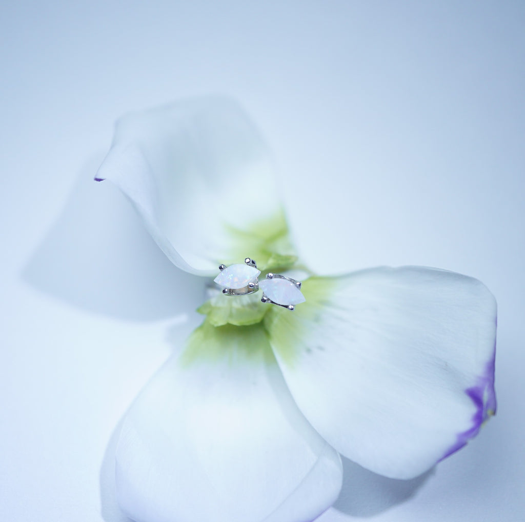 A delicate silver ring with a white marquise-cut gemstone and small clear accent stones, delicately resting on the center of a white flower with subtle green and purple accents, set against a soft light blue background.