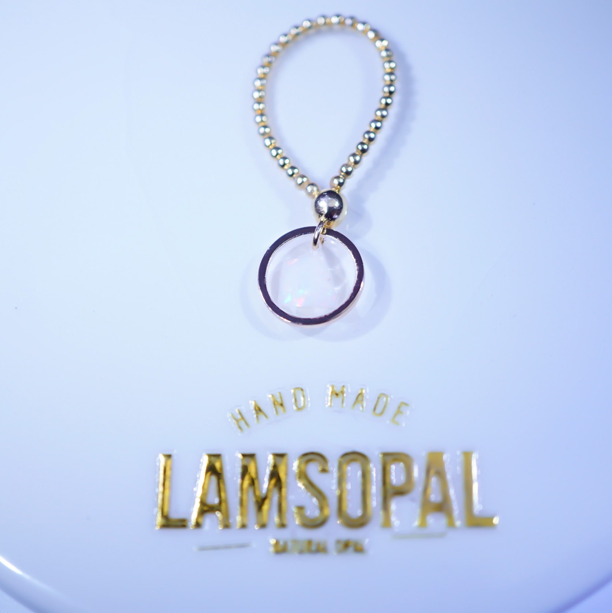 A gold beaded keychain or bag charm with a circular ring pendant, displayed above a branded gold 'LAMSOPAL HAND MADE' label on a light blue background.