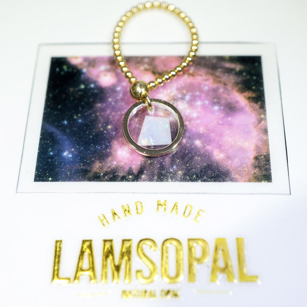 A gold beaded keychain with a clear glass disc pendant containing a white crystal, displayed on a galaxy-themed backing card with the brand name 'Lamsopal' printed below.