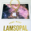 A gold beaded bracelet with a circular gold ring and a faceted iridescent white gemstone pendant, displayed on top of a galaxy print card with 'HAND MADE LAMSOPAL' text below.