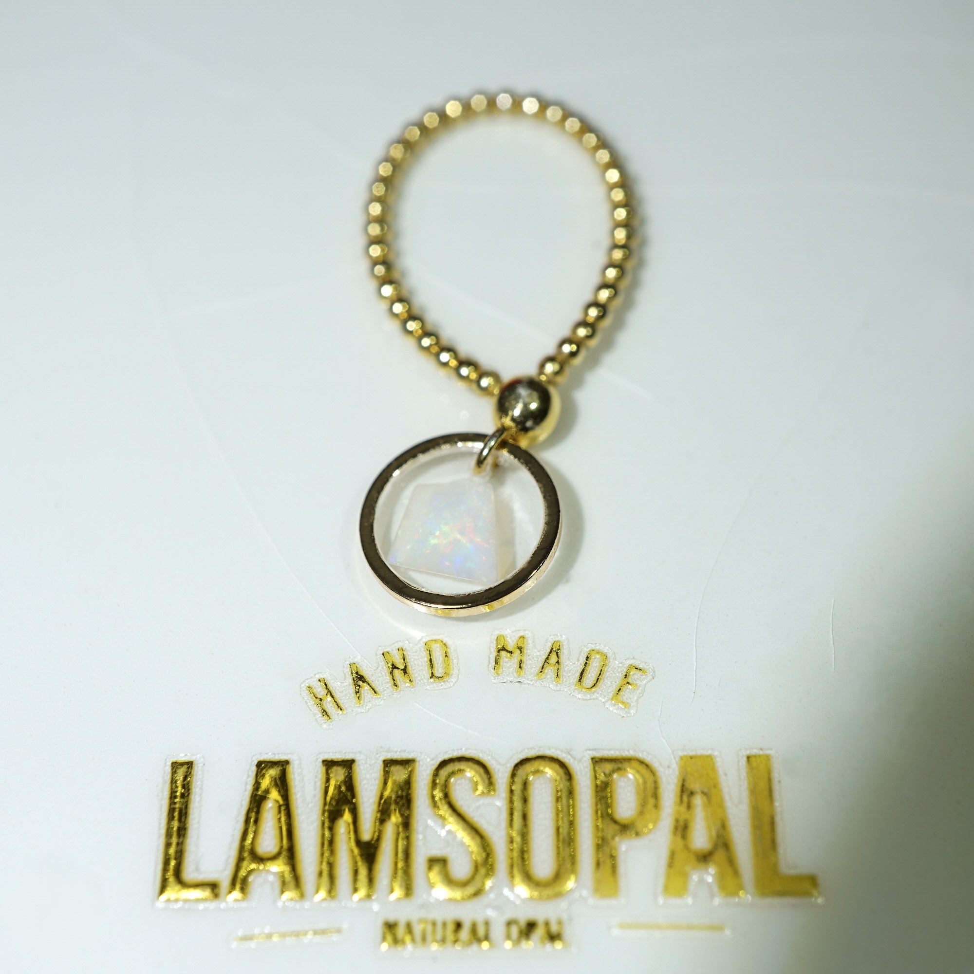 A gold-toned beaded key ring charm with a circular iridescent opal-like pendant, shown above a white card printed with the brand name Lamsopal and the words 'Hand Made' and 'Natural Opal'.