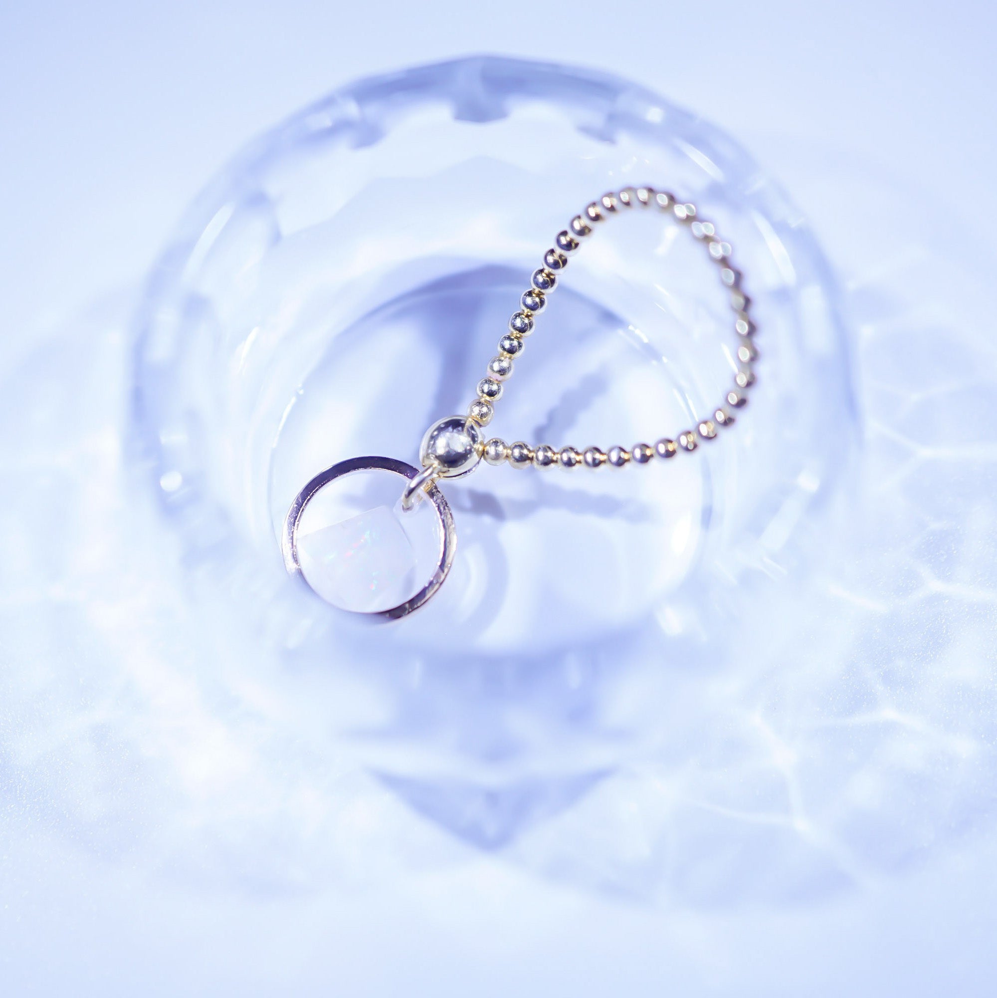 A silver-toned ball chain keychain with a thin circular ring charm, resting on a clear glass dish against a light blue background.