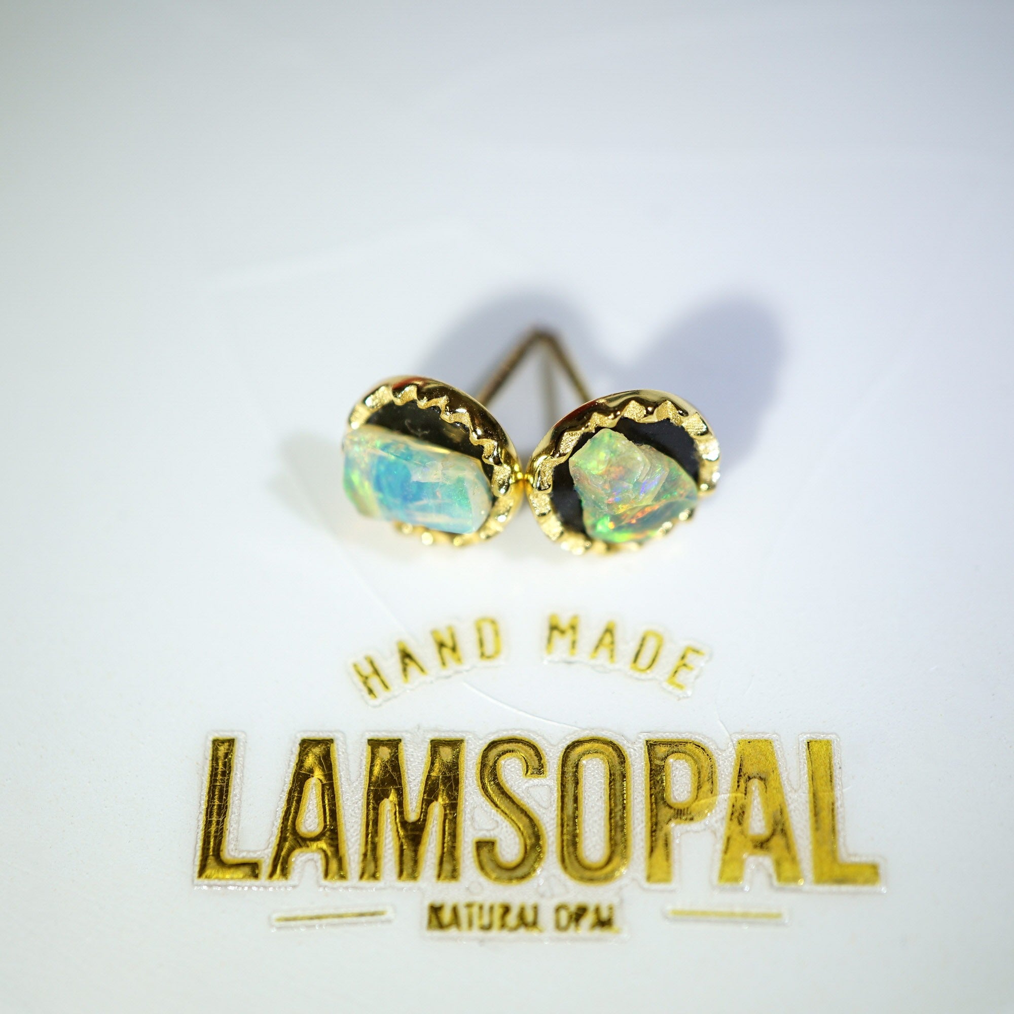 A pair of gold stud earrings with iridescent natural opal gemstones, displayed above a gold foil card that reads 'HAND MADE LAMSOPAL NATURAL OPAL'.