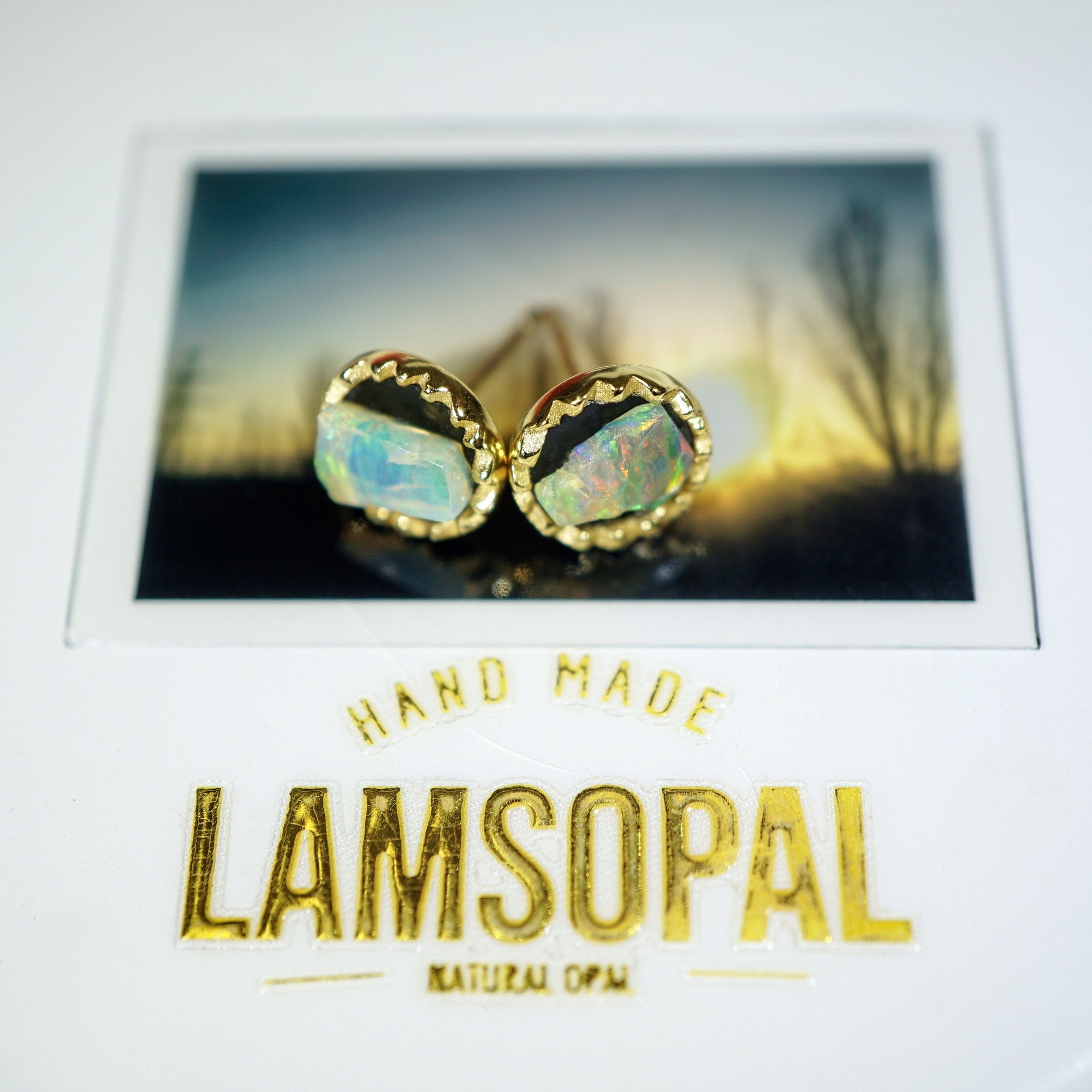 A pair of gold stud earrings with iridescent opal gemstones, displayed on a white card with a scenic photo background and the brand name 'LAMSOPAL' printed in gold.