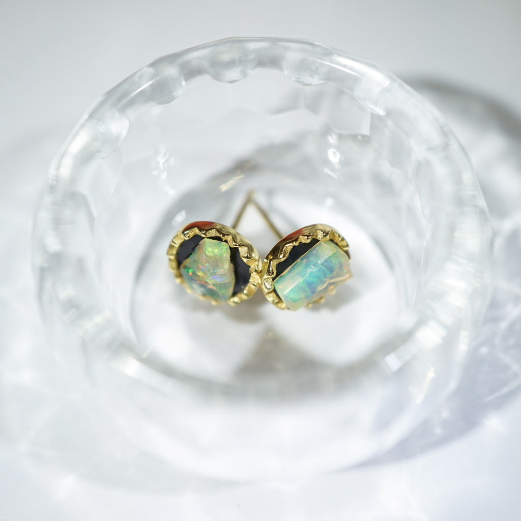 A pair of gold stud earrings with iridescent opal-like cabochons, displayed inside a clear textured glass bowl on a white surface.