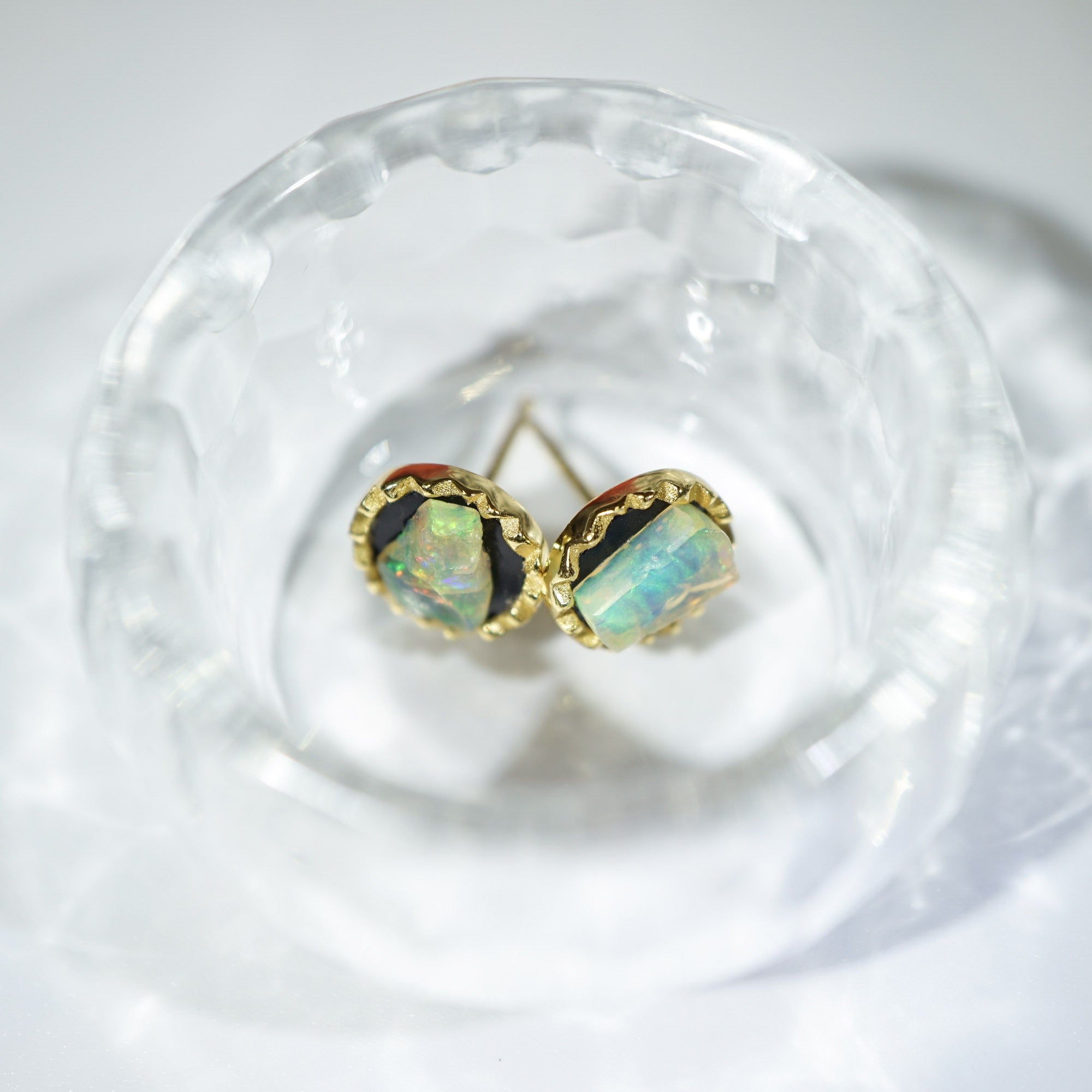 A pair of gold stud earrings with iridescent opal-like cabochons, displayed inside a clear textured glass bowl on a white surface.