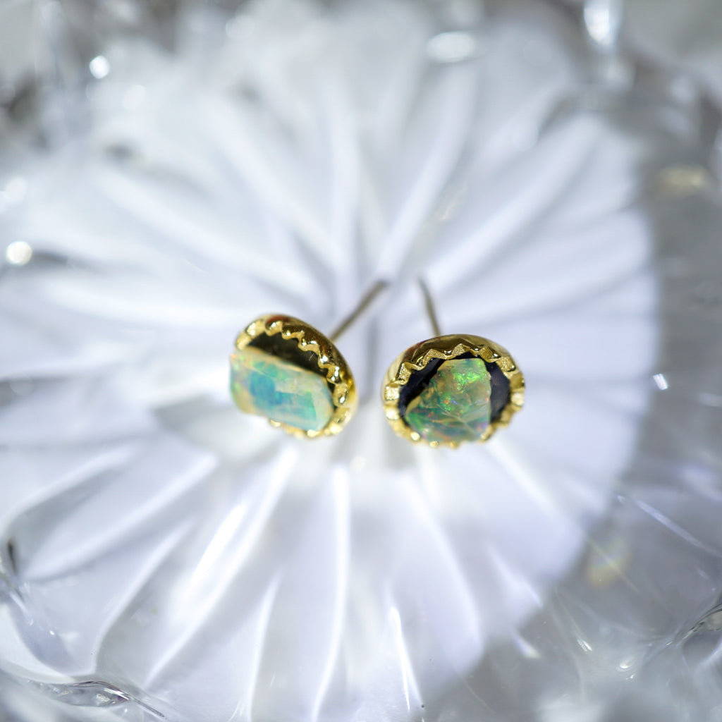 A pair of gold stud earrings with irregular, iridescent opal-like stones, displayed on a white textured glass surface.