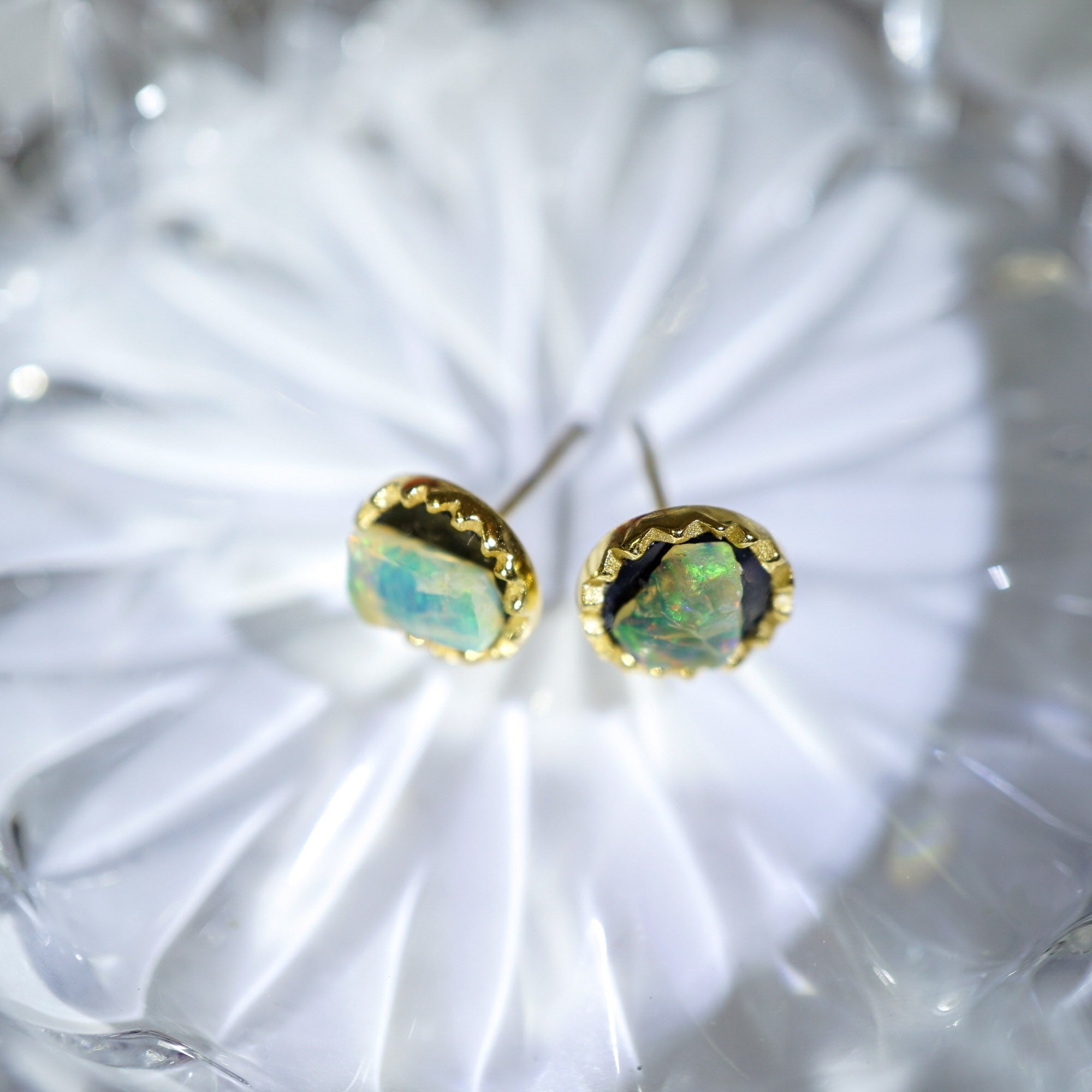 A pair of gold stud earrings with irregular, iridescent opal-like stones, displayed on a white textured glass surface.