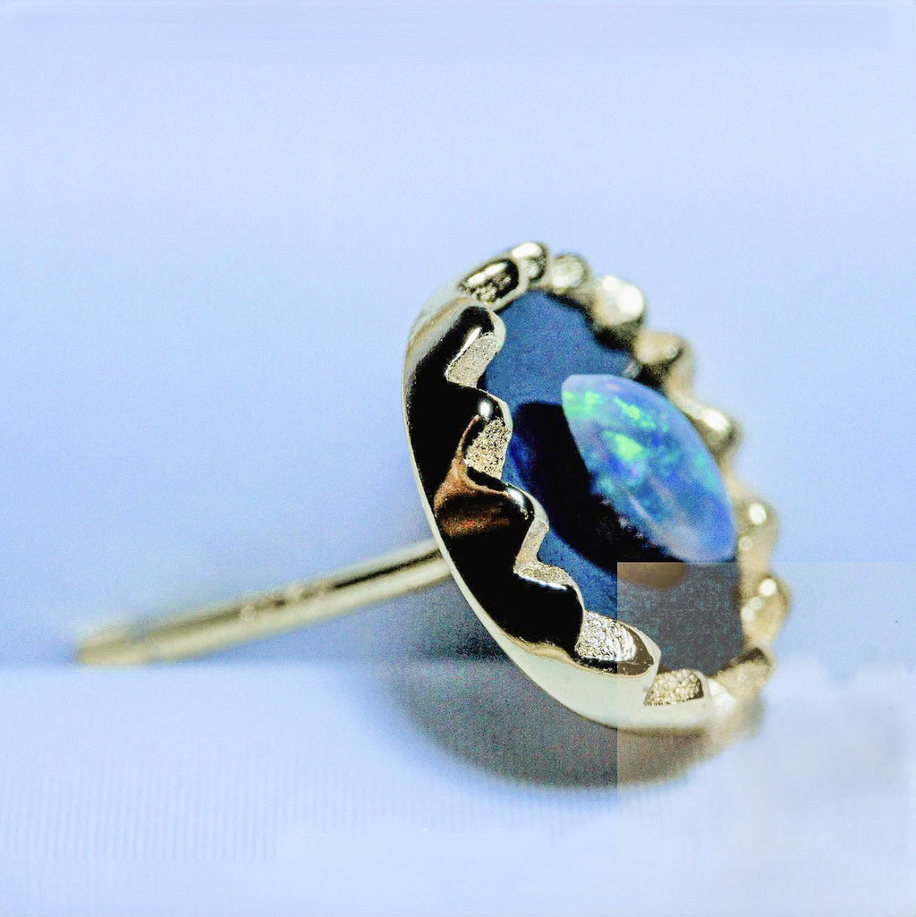 A gold ring featuring a large, irregularly shaped blue opal gemstone in a scalloped bezel setting, shown on a light blue background.