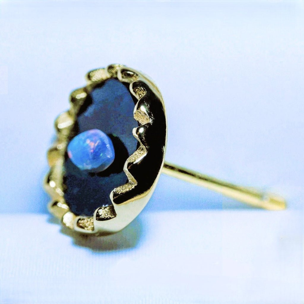 A gold stud earring featuring a large, irregularly shaped black opal with a vibrant blue center, set in a textured gold bezel with a post visible on the right.