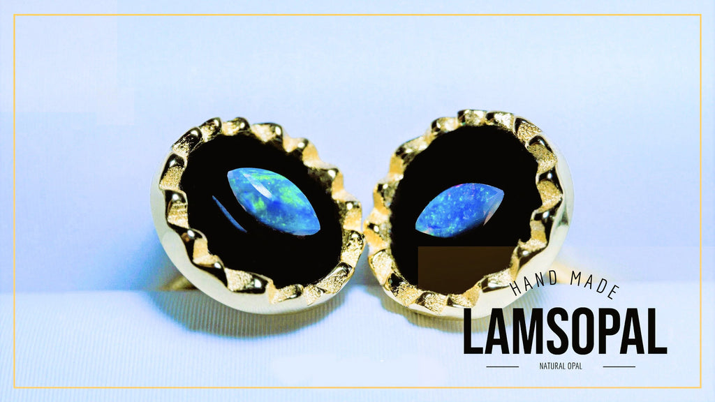 A pair of gold-toned Lamsopal stud earrings with oval-cut natural opal centers, displayed on a light blue background with a gold border. The text 'HAND MADE LAMSOPAL' is visible in the bottom right corner.