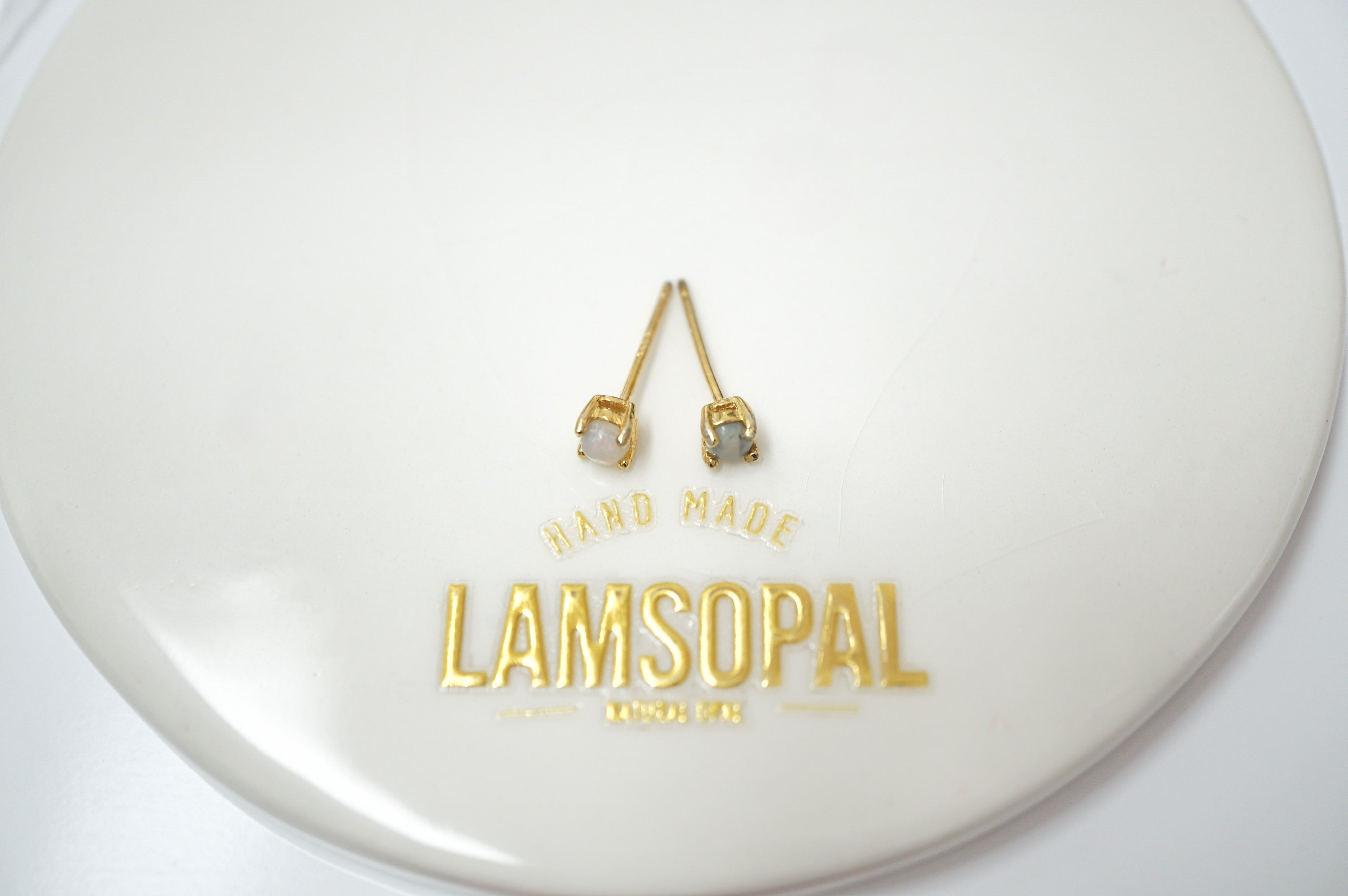 A pair of gold-toned dangle earrings with small clear stones, displayed on a glossy white circular surface with the brand name LAMSOPAL printed in gold below.