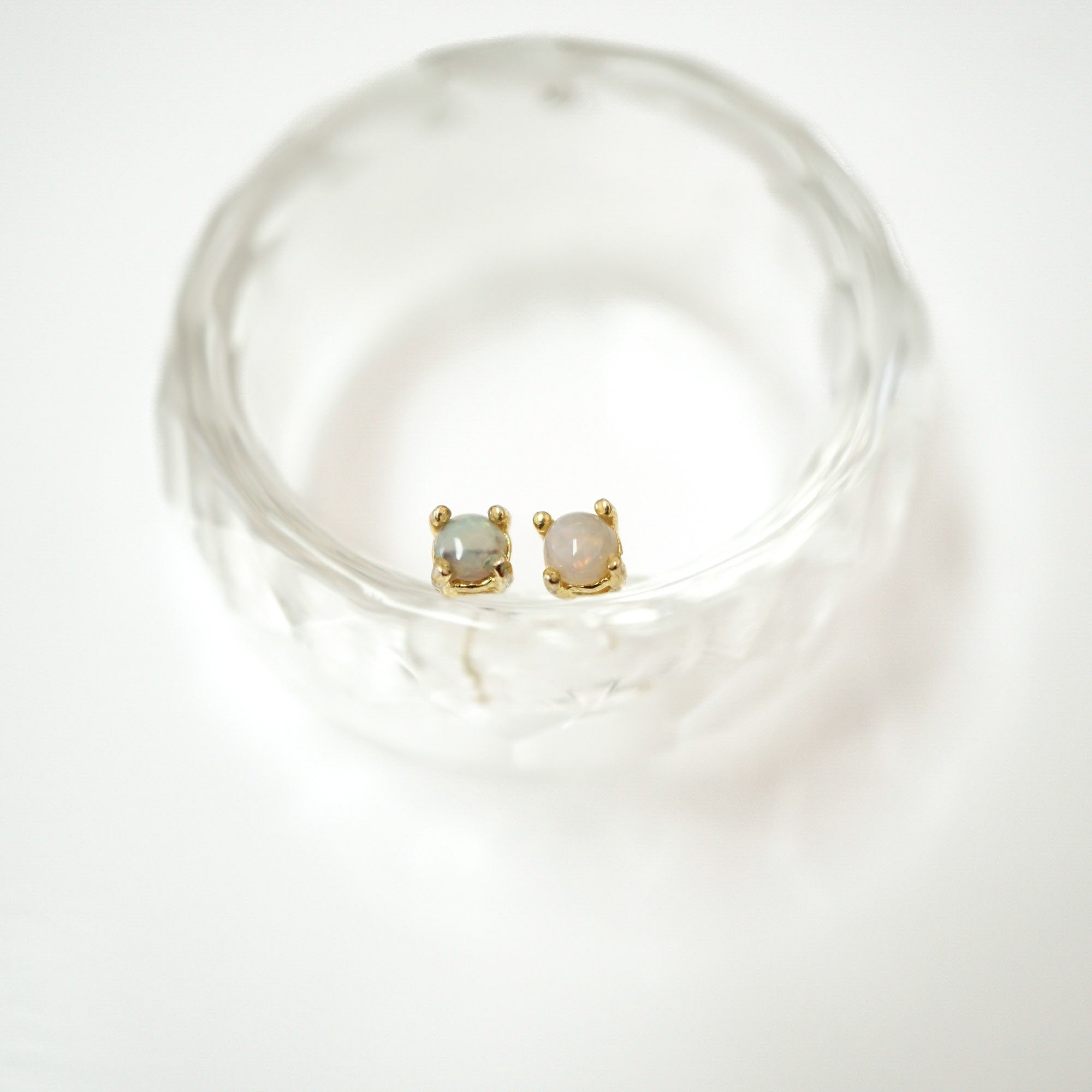 A pair of gold stud earrings with round pale gray-blue and pinkish stones, displayed inside a clear textured glass bowl on a white background.