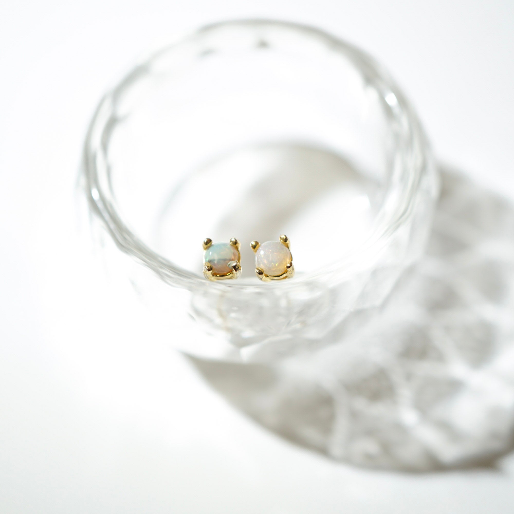 A pair of gold stud earrings with oval iridescent opal-like stones, resting inside a clear textured glass bowl on a bright white background.