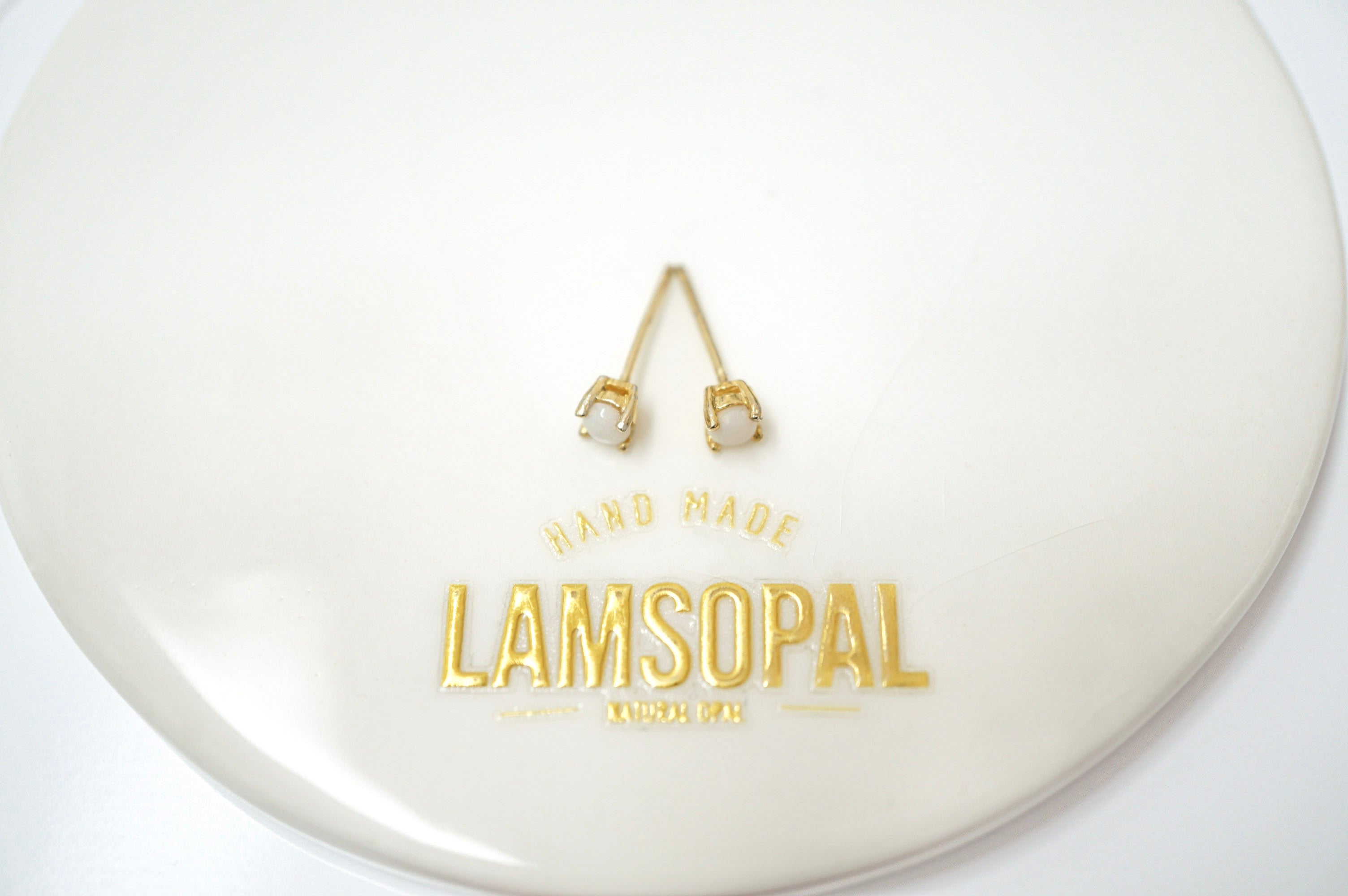A pair of small gold-toned stud earrings with clear rectangular stones, displayed on a glossy white circular surface with the brand name LAMSOPAL printed in gold below.