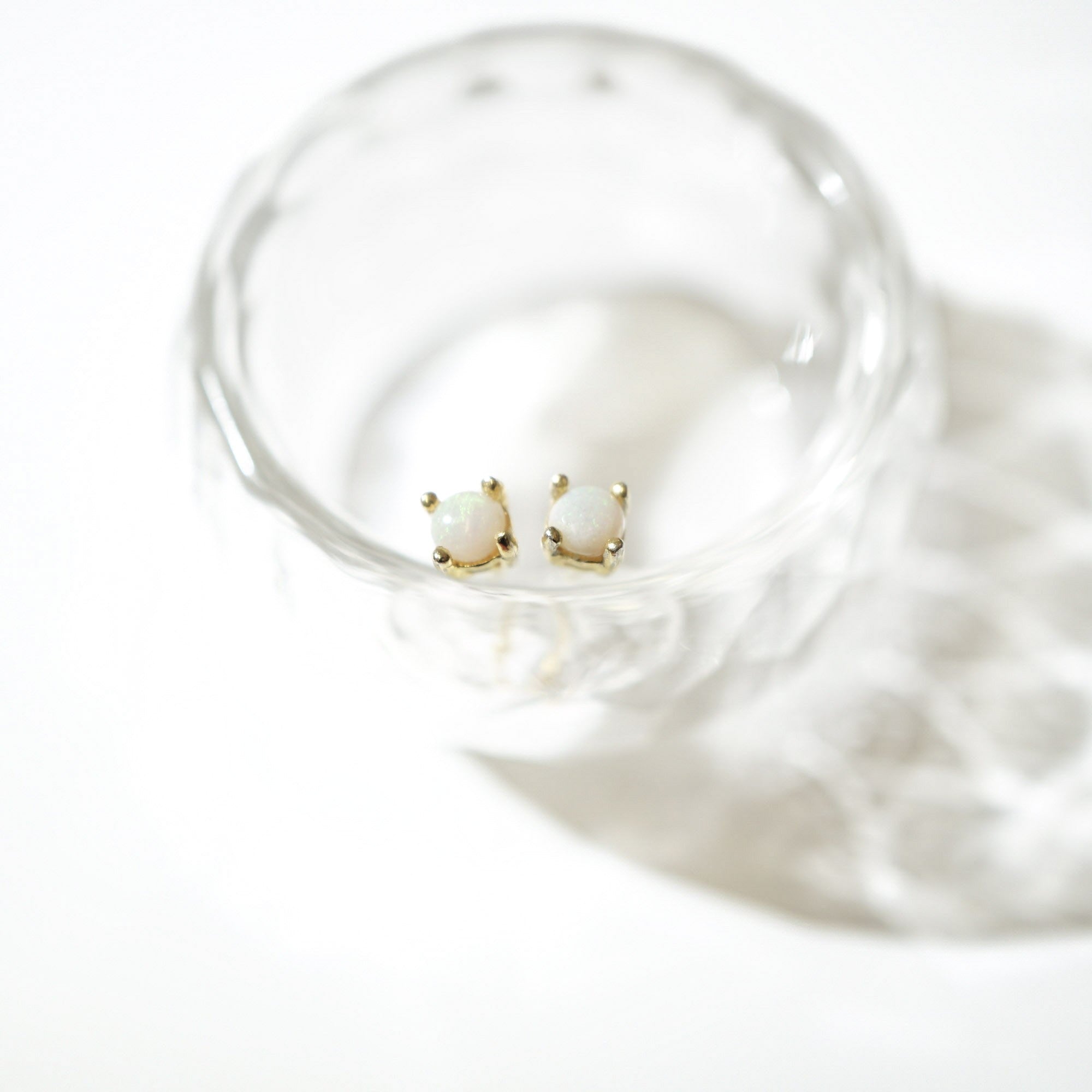 A pair of gold stud earrings with oval white stones, resting inside a clear glass dish on a bright white background.