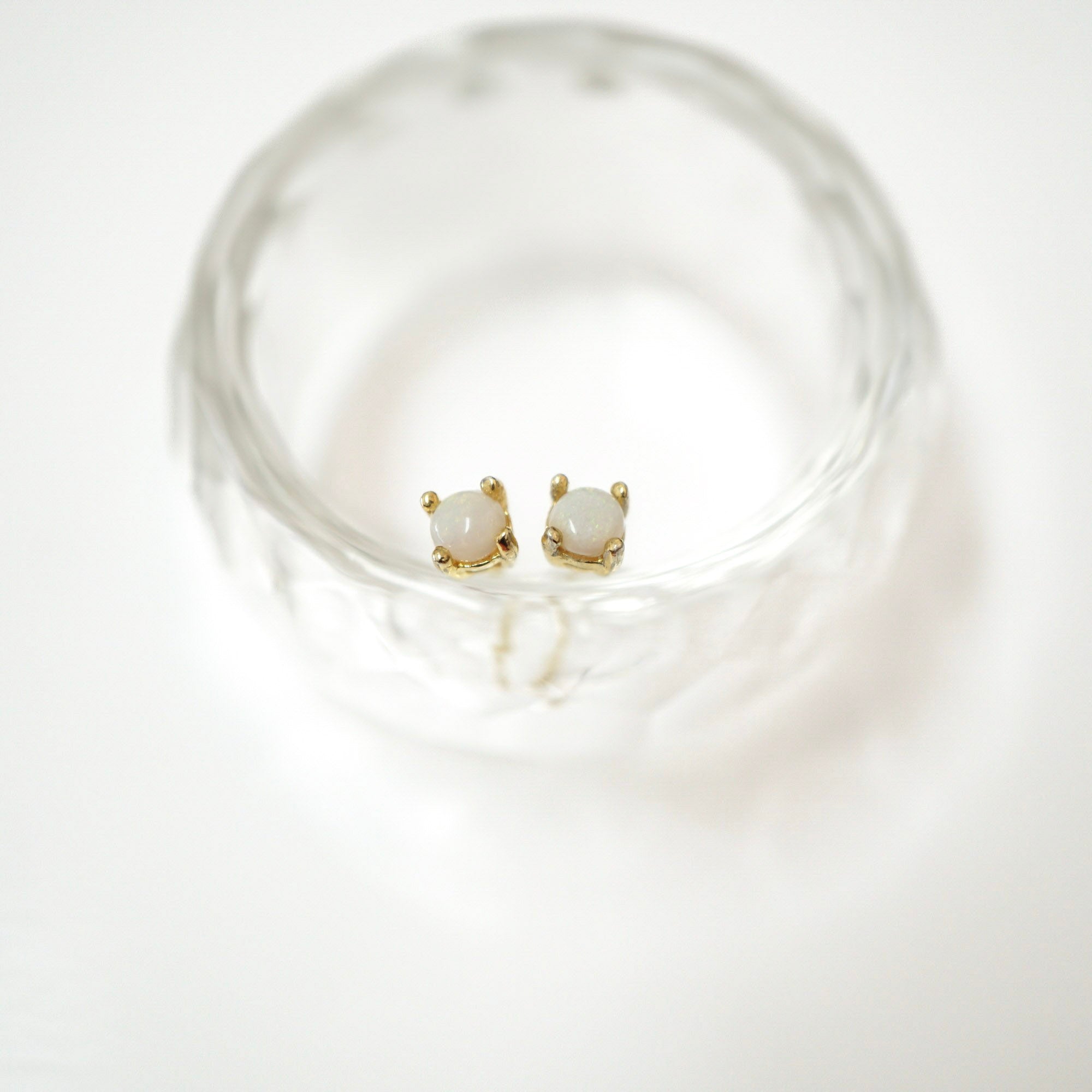 A pair of gold stud earrings with oval milky white stones, displayed inside a clear textured glass bowl on a white background.