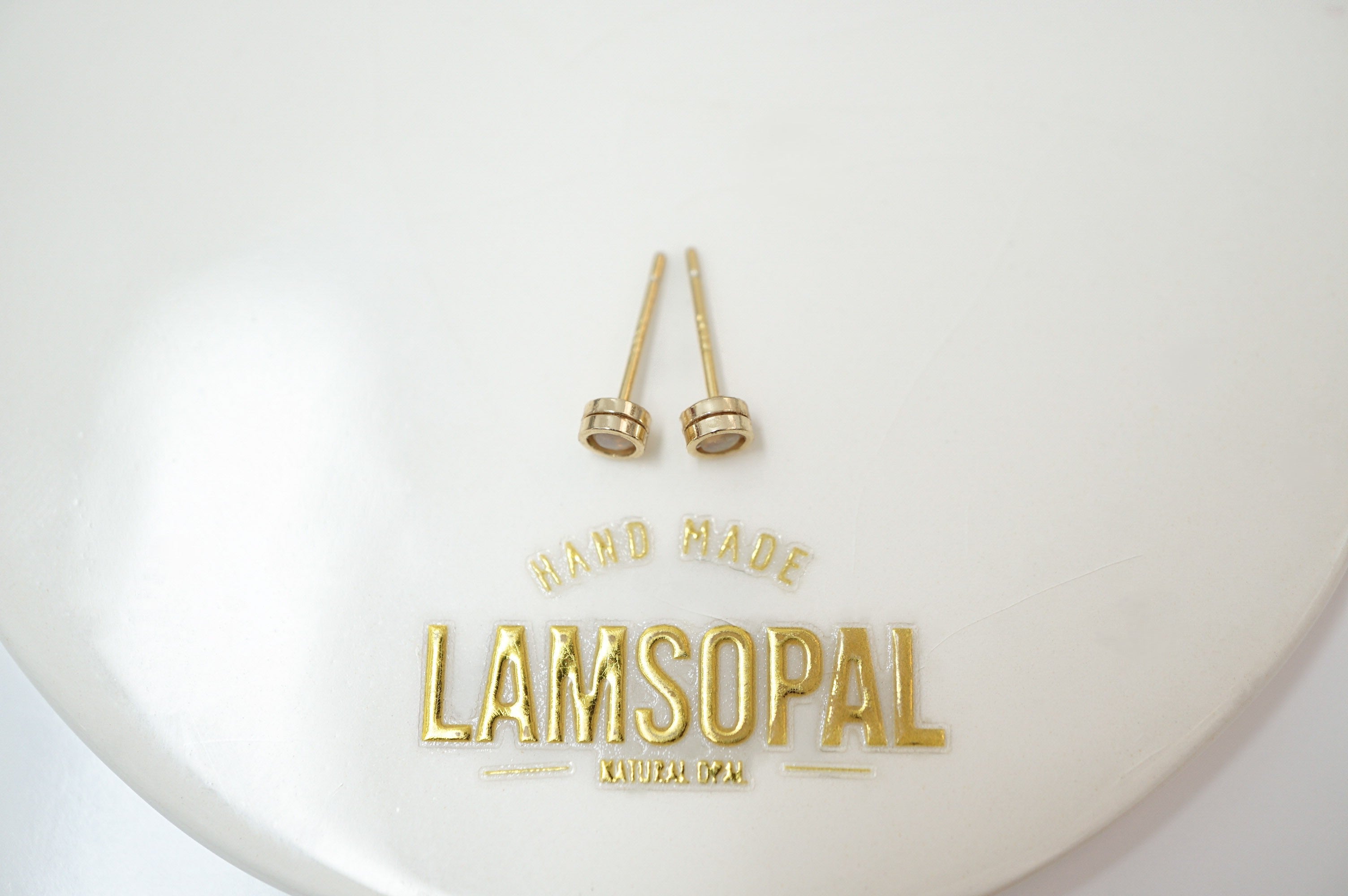 A pair of small gold-toned stud earrings displayed on a white oval surface with the brand name LAMSOPAL printed in gold below.