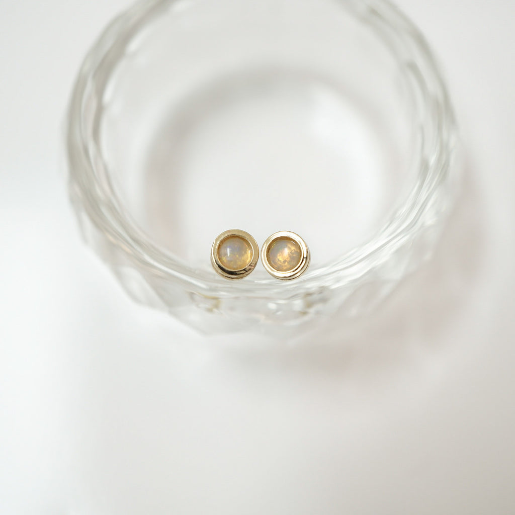 A pair of gold-toned stud earrings with oval milky white stones, displayed inside a clear faceted glass bowl on a white background.