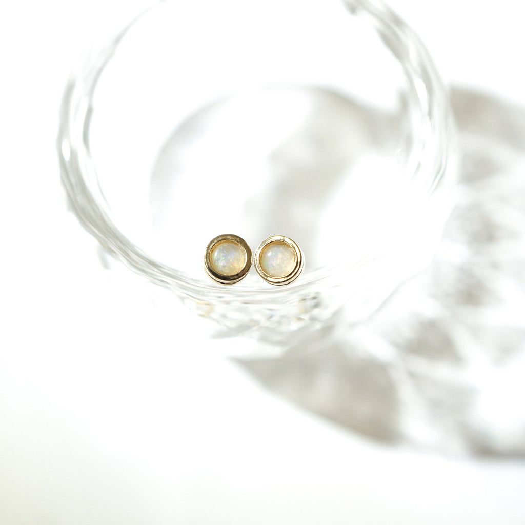 A pair of gold stud earrings with round white opal inlays, resting inside a clear glass bowl on a bright white background.