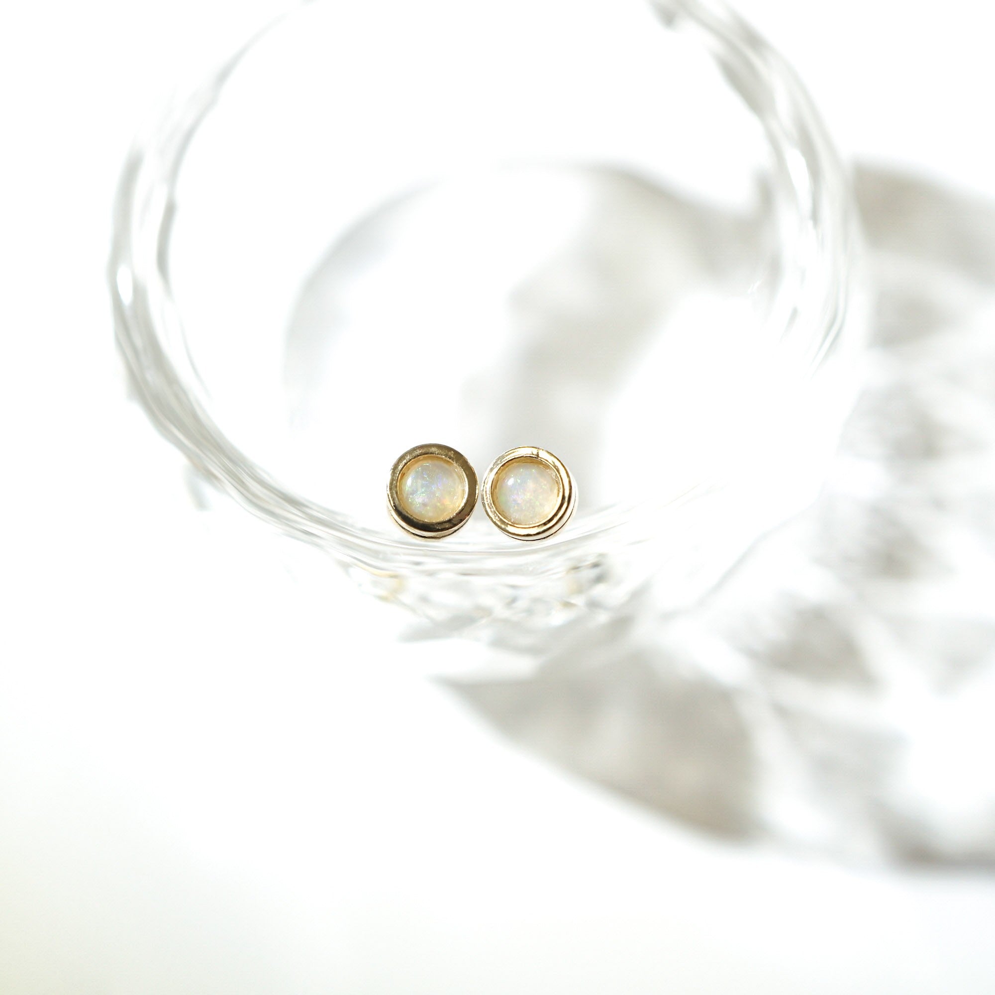 A pair of gold stud earrings with round white opal inlays, resting inside a clear glass bowl on a bright white background.
