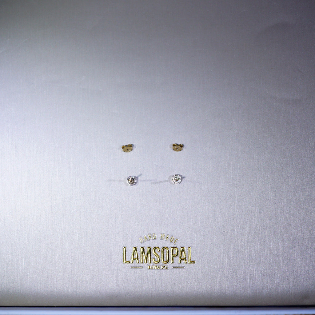 A close-up of a silver-toned jewelry box lid featuring the gold-embossed brand name 'LAMSOPAL'. On the lid, four small stud earrings are arranged in two rows: two gold-tone crescent moon studs and two silver-tone round stud earrings with clear stones.