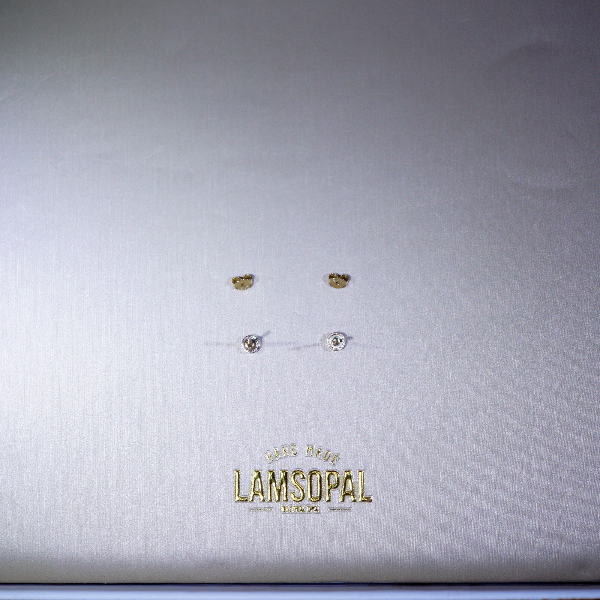 A close-up of a silver-toned jewelry box lid featuring the gold-embossed brand name 'LAMSOPAL'. On the lid, four small stud earrings are arranged in two rows: two gold-tone crescent moon studs and two silver-tone round stud earrings with clear stones.