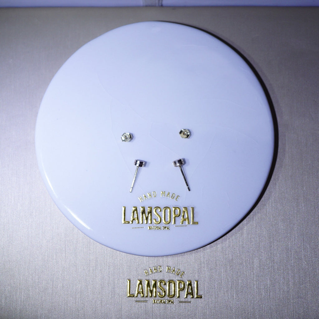 A pair of small silver stud earrings displayed on a round white backing card with gold Lamsopal branding, set against a brushed metallic gray background.