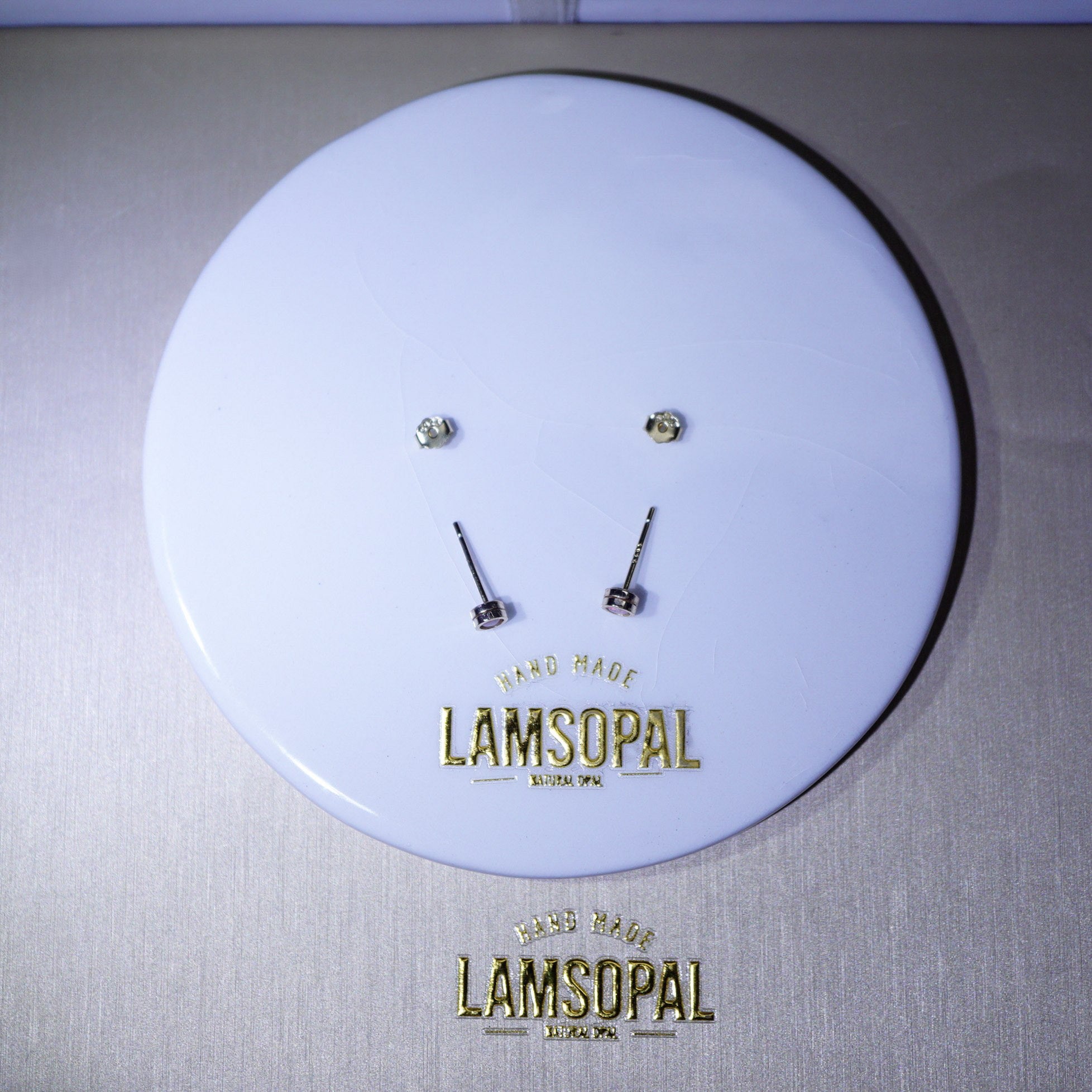 A pair of small gold-toned stud earrings displayed on a round white branded backing card. The card shows the brand name Lamsopal in gold lettering and reads 'Hand Made Lamsopal London.