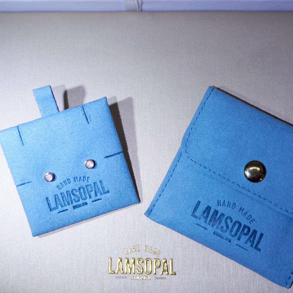 Two light blue suede-like Lamsopal card cases on a light gray textured background, one flat square case with two metal studs and one triangular flap wallet with a snap button. A gold Lamsopal logo is visible at the bottom.