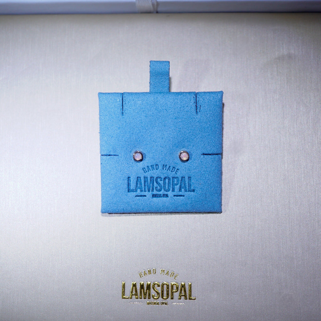 A small blue square jewelry box or packaging piece with a tab at the top, featuring the Lamsopal brand name and two small circular studs, shown on a light textured background with a larger gold-toned Lamsopal logo below.