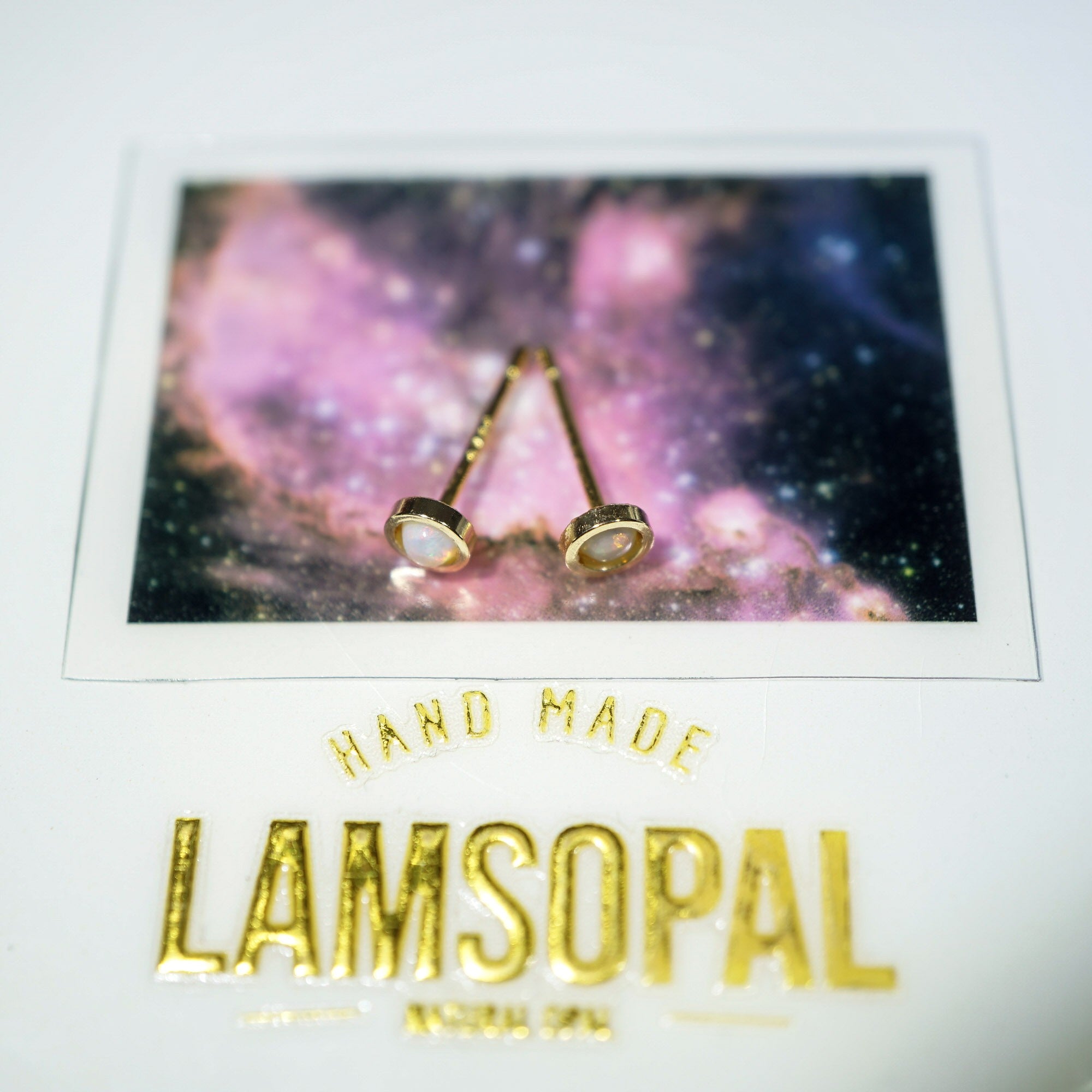 A pair of gold post earrings with oval clear stones displayed on a printed card featuring a pink galaxy background and the brand name Lamsopal.