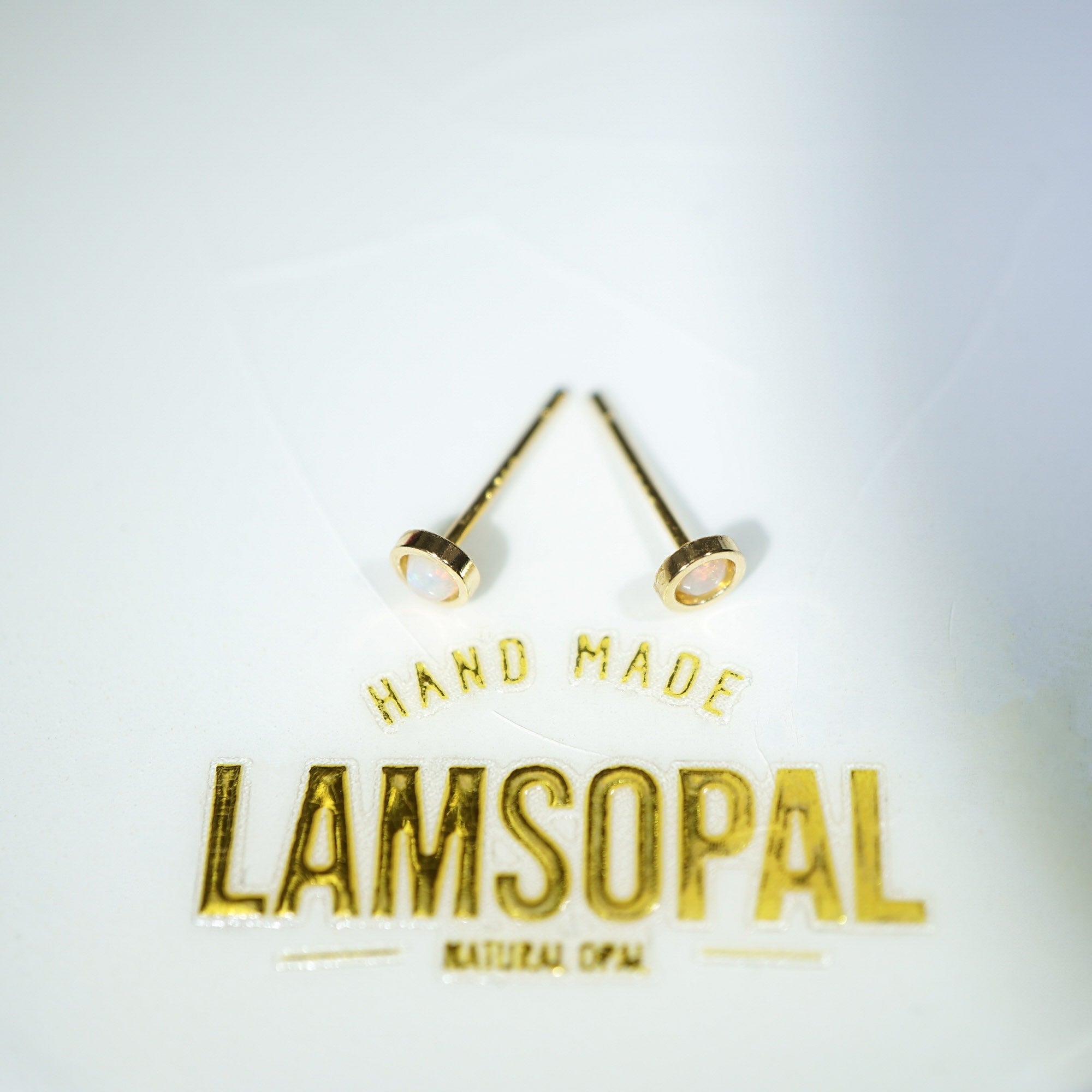 A pair of gold-colored post earrings with circular disc studs, displayed on a white background above the gold foil text 'LAMSOPAL' and 'HAND MADE'.