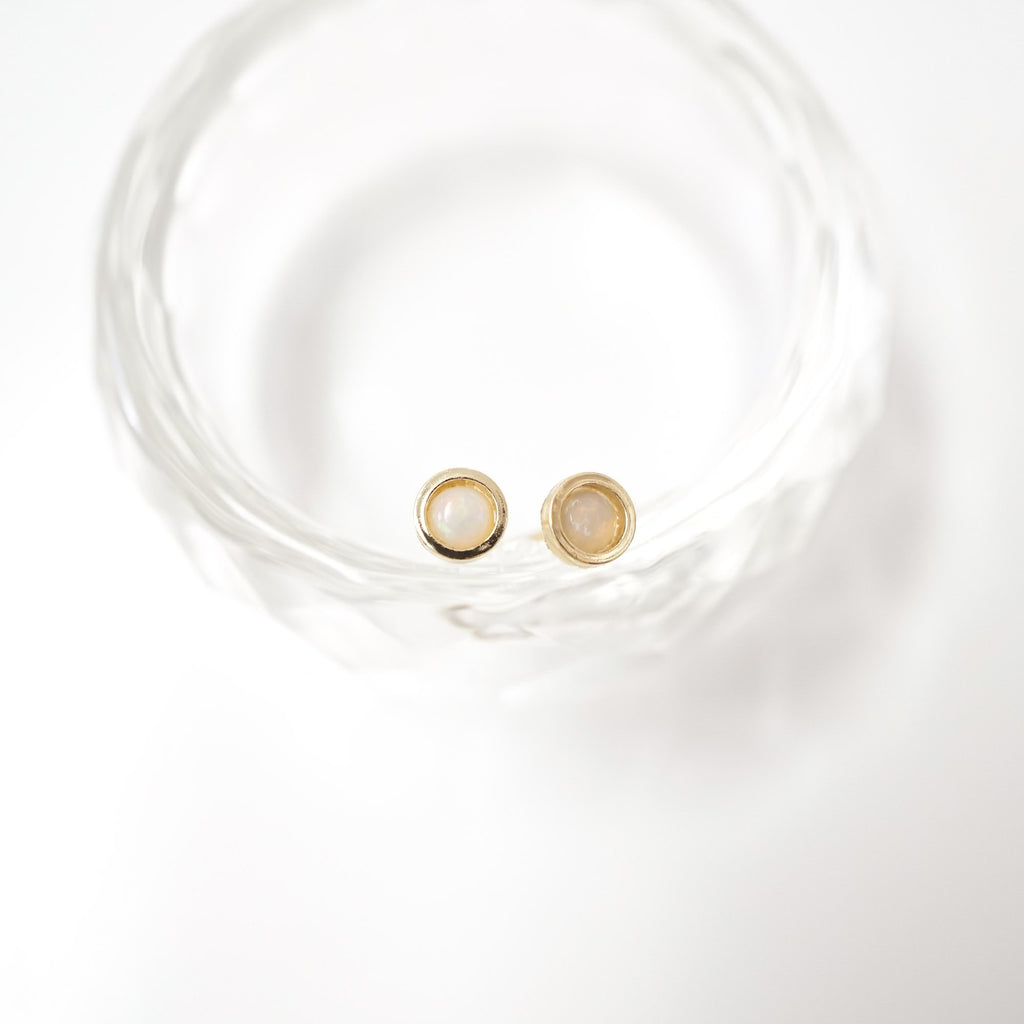 A pair of gold stud earrings with oval milky white inlays, resting inside a clear textured glass bowl on a white background.