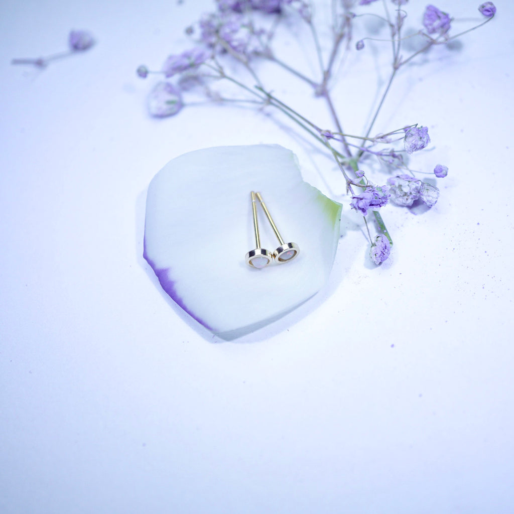 A pair of gold-toned stud earrings with small white oval inlays, displayed on a white petal-shaped coaster with dried lavender flowers around them on a pale background.