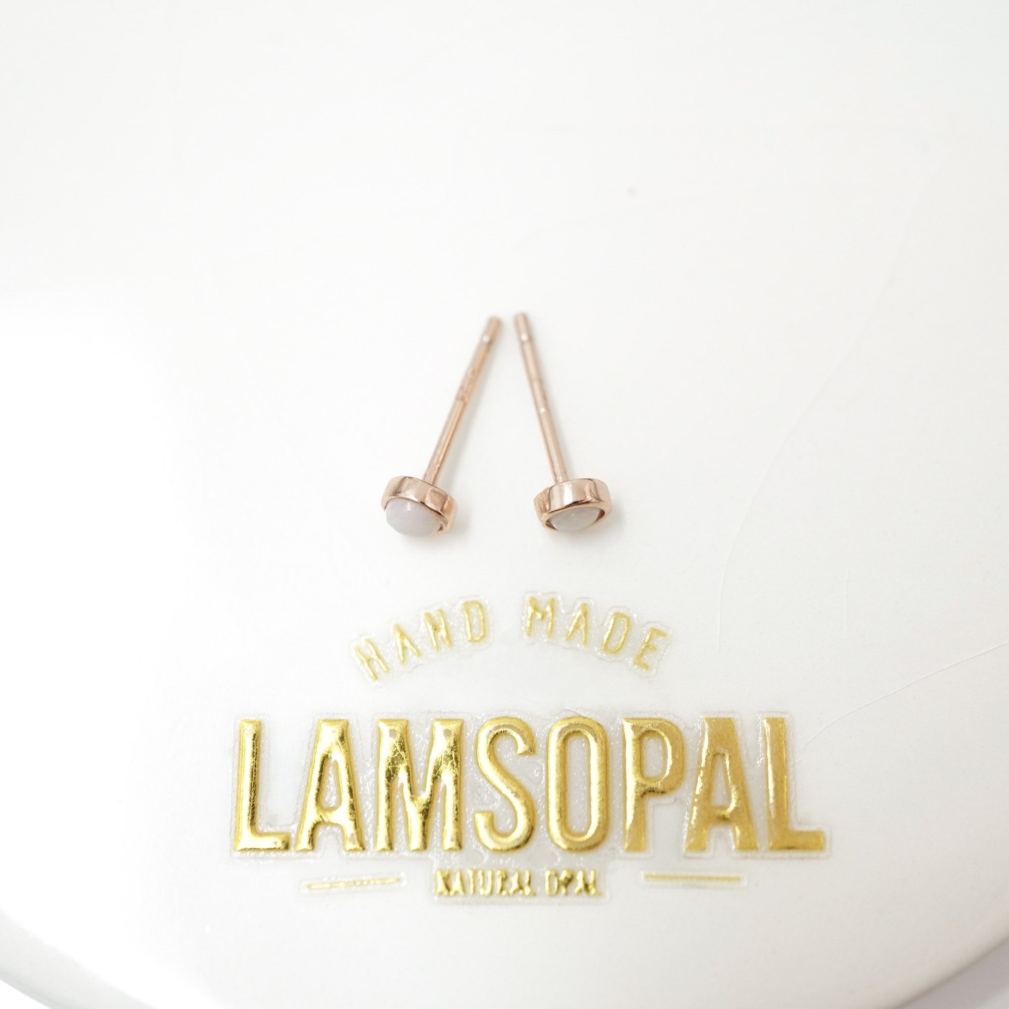 A pair of rose gold post earrings with oval white cabochon stones, displayed on a white background above gold foil branding that reads 'HAND MADE LAMSOPAL'.