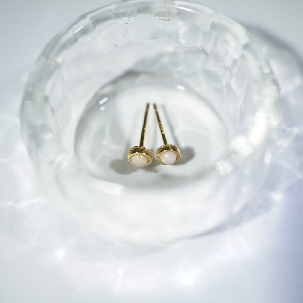 A pair of gold stud earrings with oval white pearl-like centers, displayed inside a clear faceted glass dish on a soft white surface.