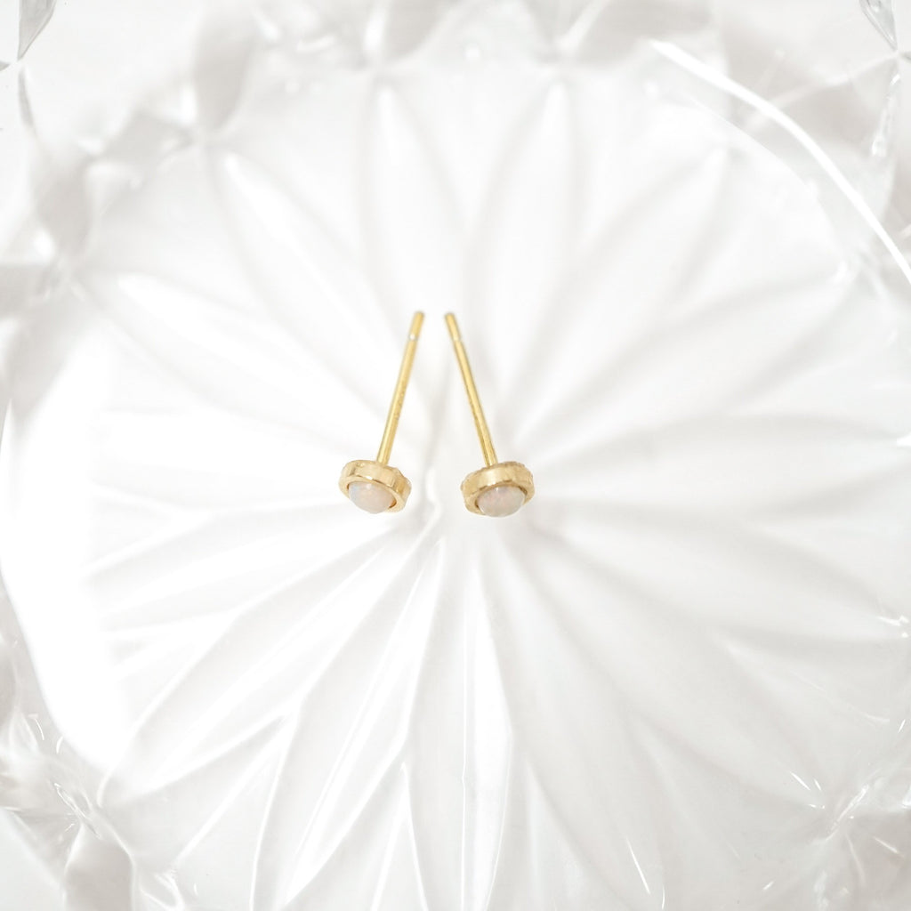 A pair of gold post earrings with oval cream-colored stone settings, displayed on a white textured glass plate.