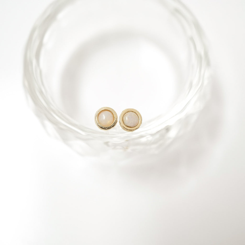 A pair of gold stud earrings with oval milky white stone centers, resting inside a clear textured glass bowl on a white background.