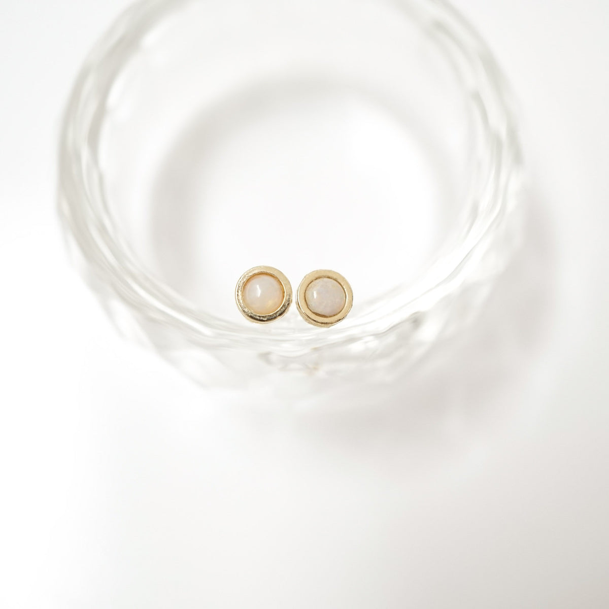 A pair of gold stud earrings with oval milky white stone centers, resting inside a clear textured glass bowl on a white background.