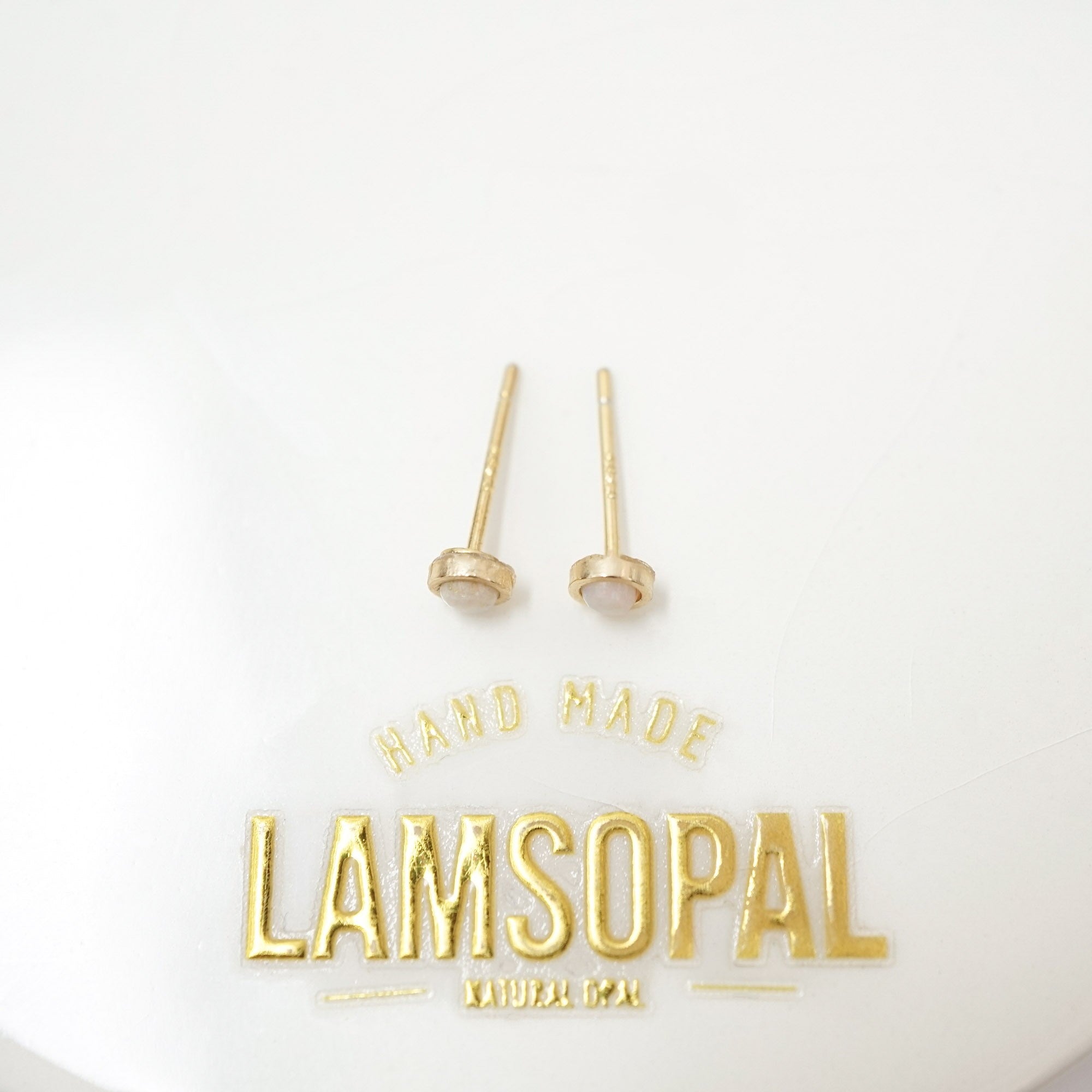 A pair of gold post earrings with oval bezel-set pale pink stones, displayed on a white surface above a gold 'HAND MADE LAMSOPAL' logo.