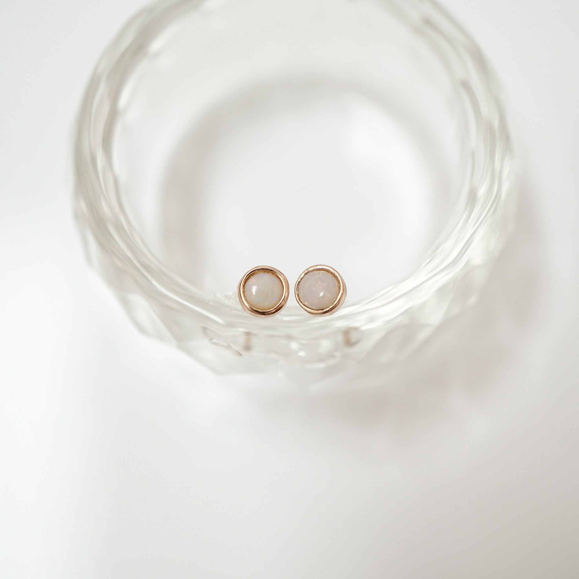 A pair of rose gold stud earrings with oval milky white gemstones, resting inside a clear textured glass bowl on a white surface.