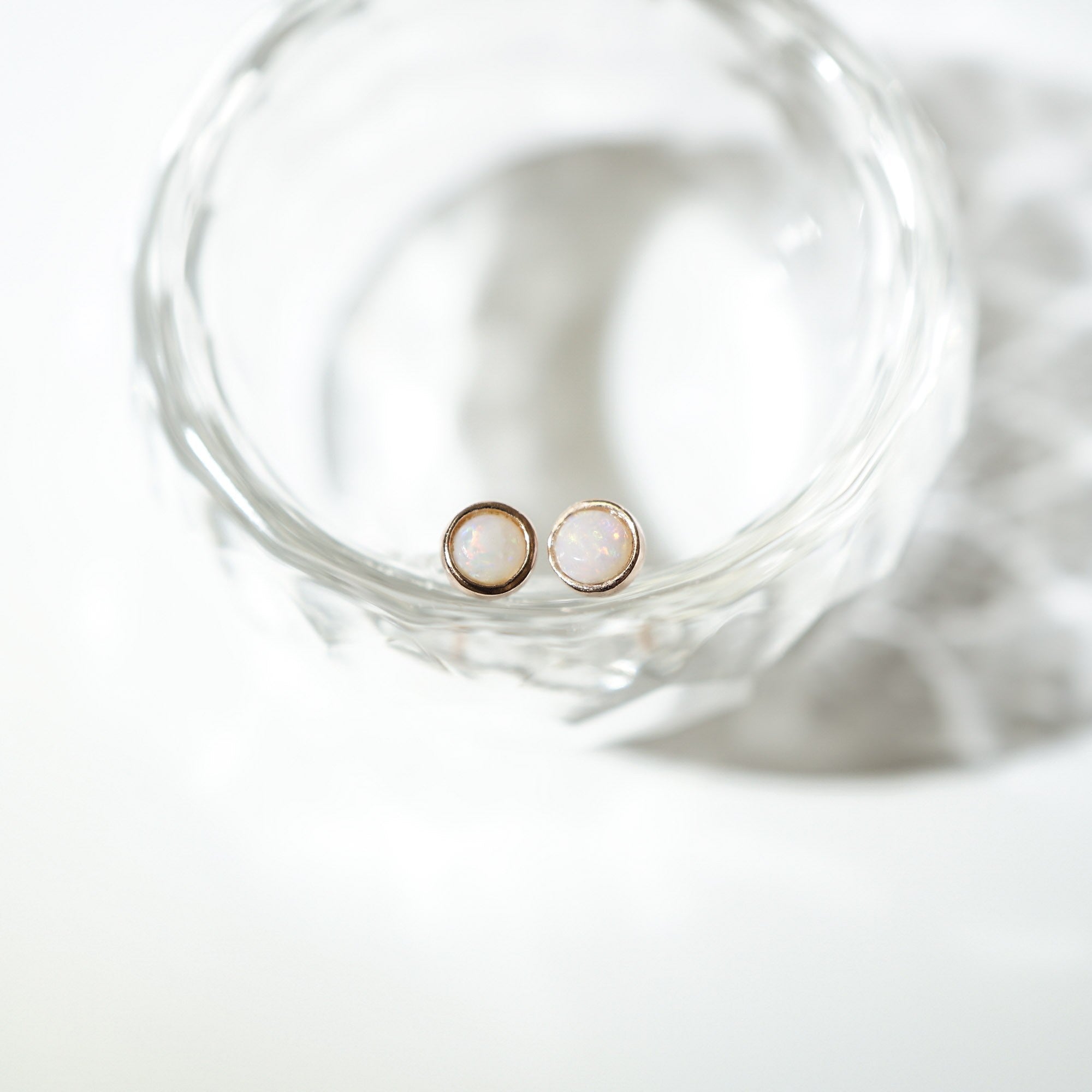 A pair of small round stud earrings with pale iridescent centers and gold-toned edges, resting inside a clear glass dish on a bright white surface.
