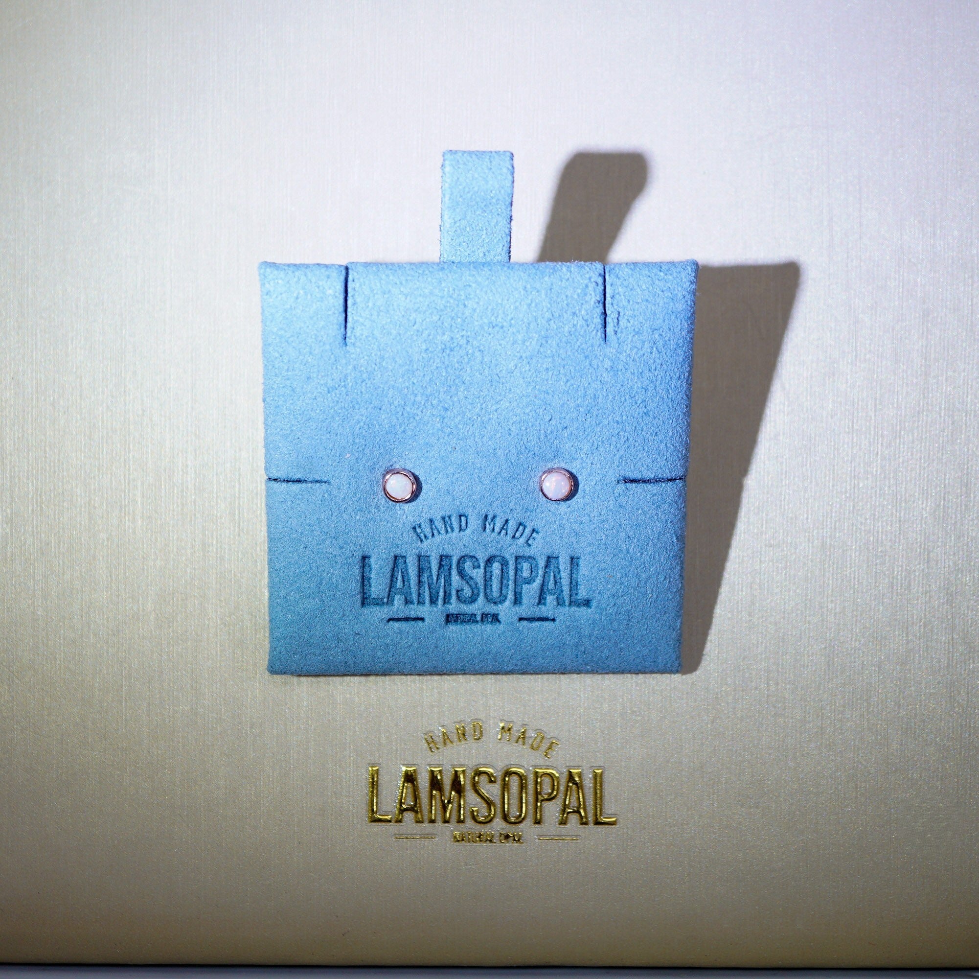 A light blue square Lamsopal hand-made leather patch with a small tab at the top and two tiny pink eye details, displayed on a textured beige background with the Lamsopal logo printed below.