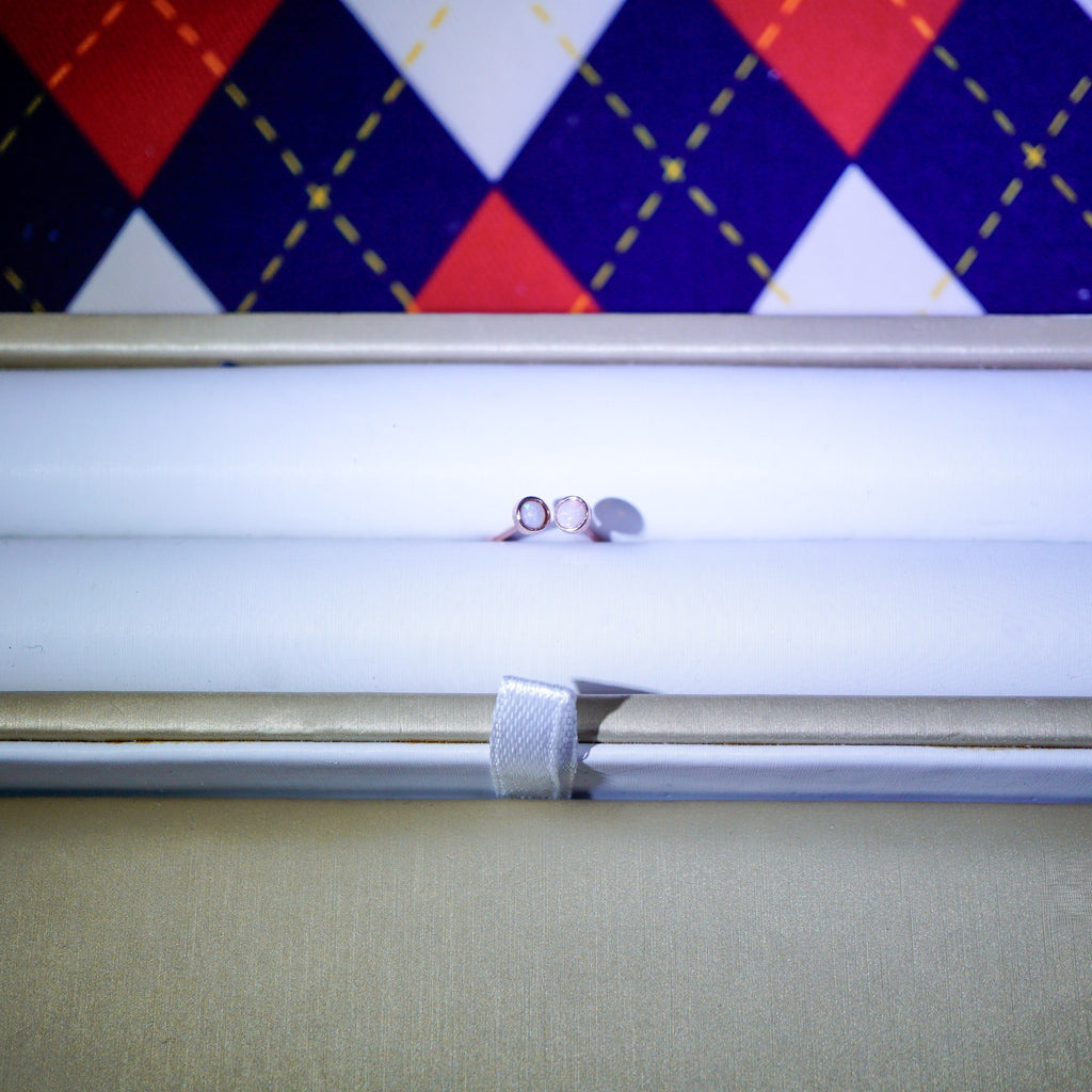 A pair of small silver hoop earrings displayed horizontally inside an open white jewelry box, with a colorful argyle-patterned fabric background visible above.
